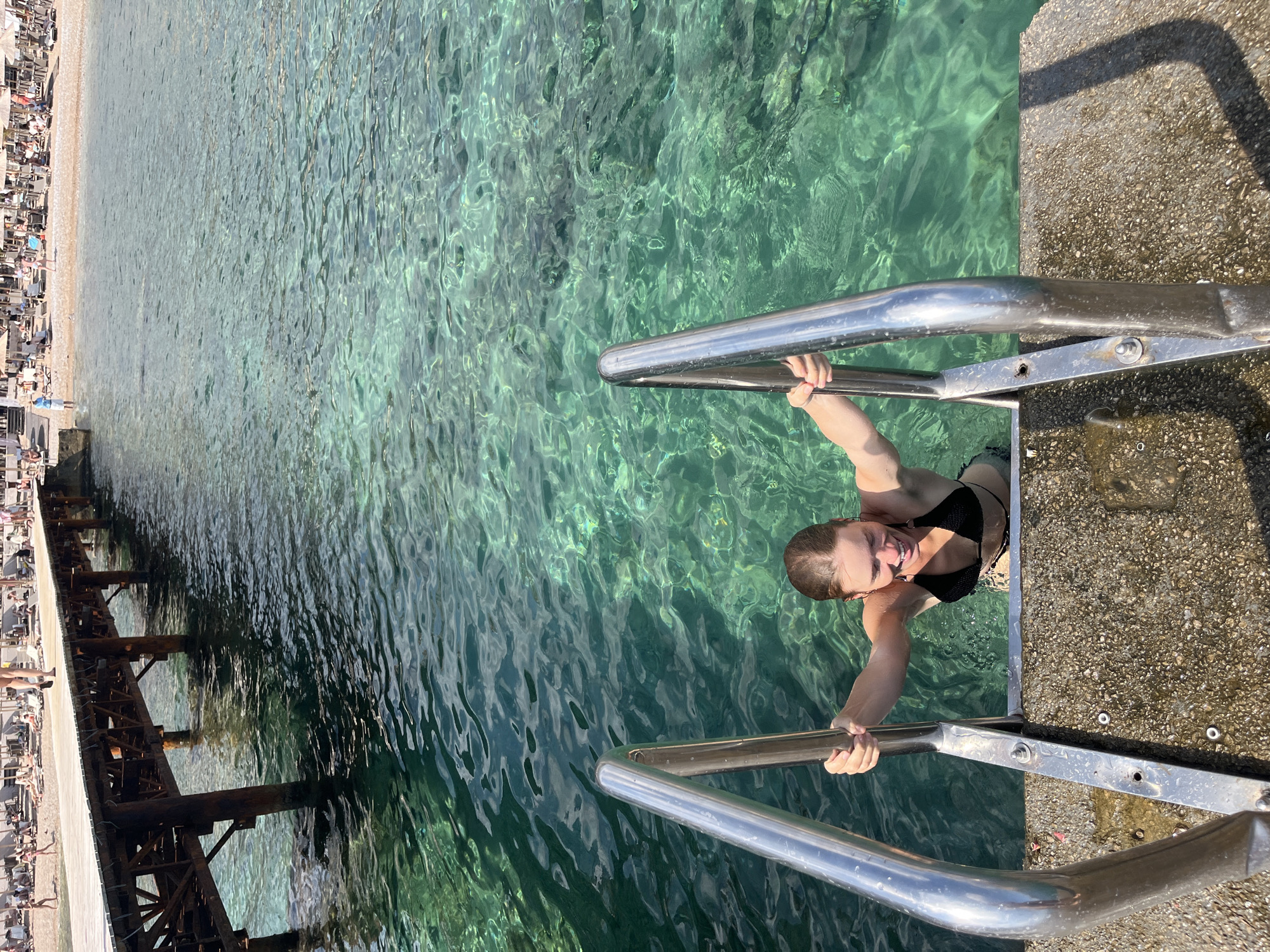 A young woman wearing a black bikini emerges from clear, turquoise water using a stainless steel ladder attached to a concrete dock. She is smiling and holding onto the ladder's handrails. The water is transparent, revealing the seabed. In the background, a wooden pier extends into the water, and a sandy beach with numerous lounge chairs and umbrellas is visible, with several people relaxing. The overall setting suggests a sunny day at a coastal resort. The location is likely near the old town of Budva, Montenegro, given the presence of a pier and beach with resort amenities.