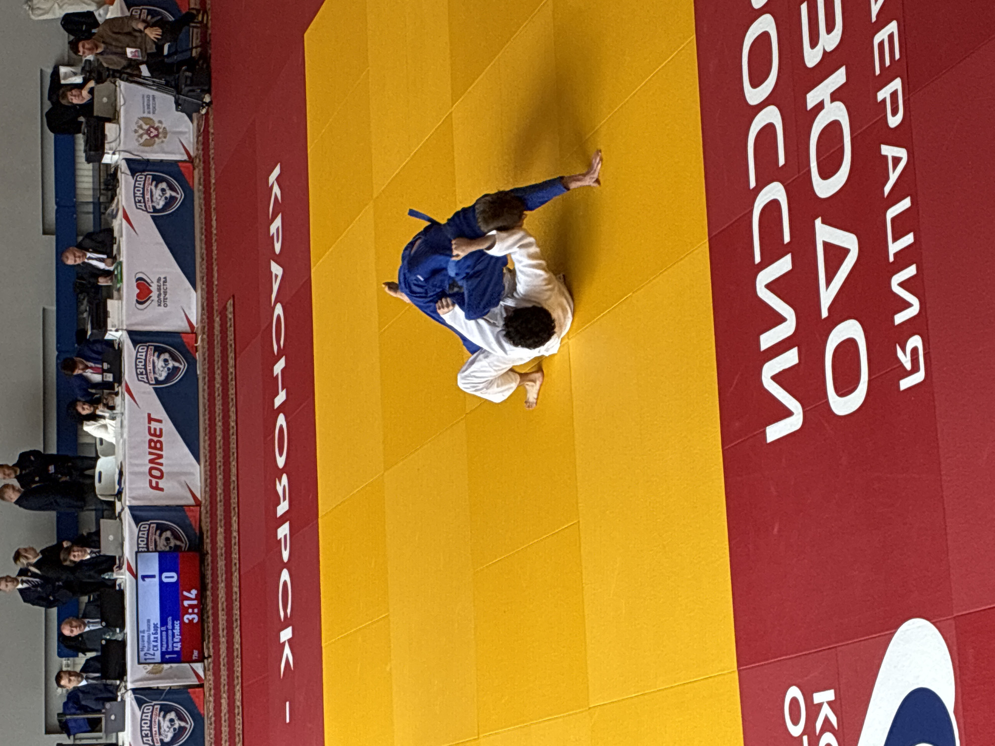 Here is a forensic description of the image:
Two competitors, one wearing a blue judogi and the other a white judogi, are grappling on a yellow and red judo mat. The mat is surrounded by advertisements and promotional materials in Russian script. The text "КРАСНОЯРСК" is prominently displayed, indicating the location of the event. A digital scoreboard shows the score as 1 to 0 with a time of 3:14. Spectators are seated in the background. The event appears to be a judo tournament in Krasnoyarsk.