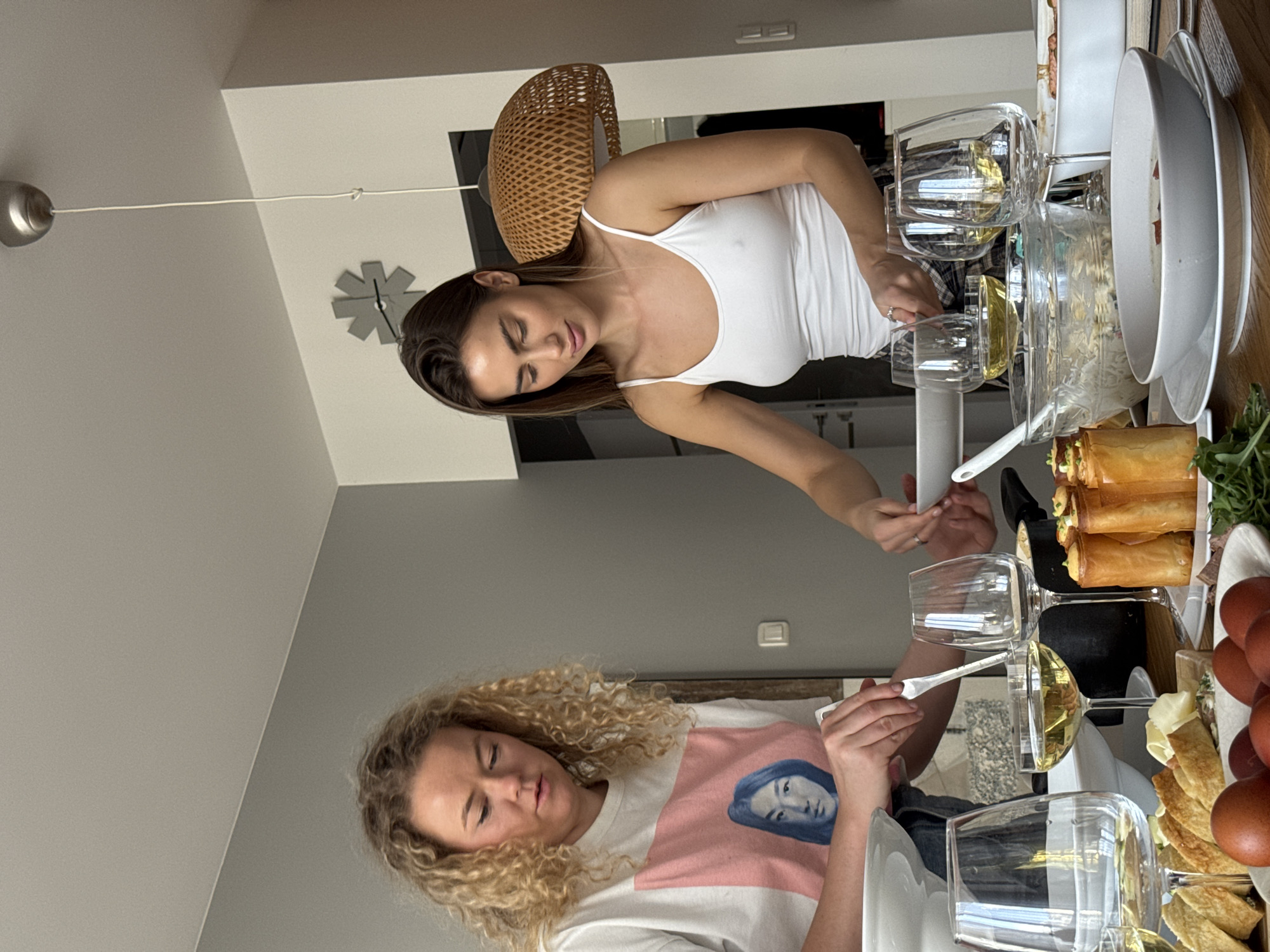 Two women are serving food at a table. One woman has brown hair and is wearing a white tank top. The other woman has curly blonde hair and is wearing a white shirt with a pink stripe and a blue image on it. They are standing next to a table filled with food, including phyllo pastries, eggs, and a salad. There are also wine glasses on the table. There is a lamp and a clock in the background. The location appears to be inside a home. I am unable to confirm the location in the city of Warsaw, Poland.