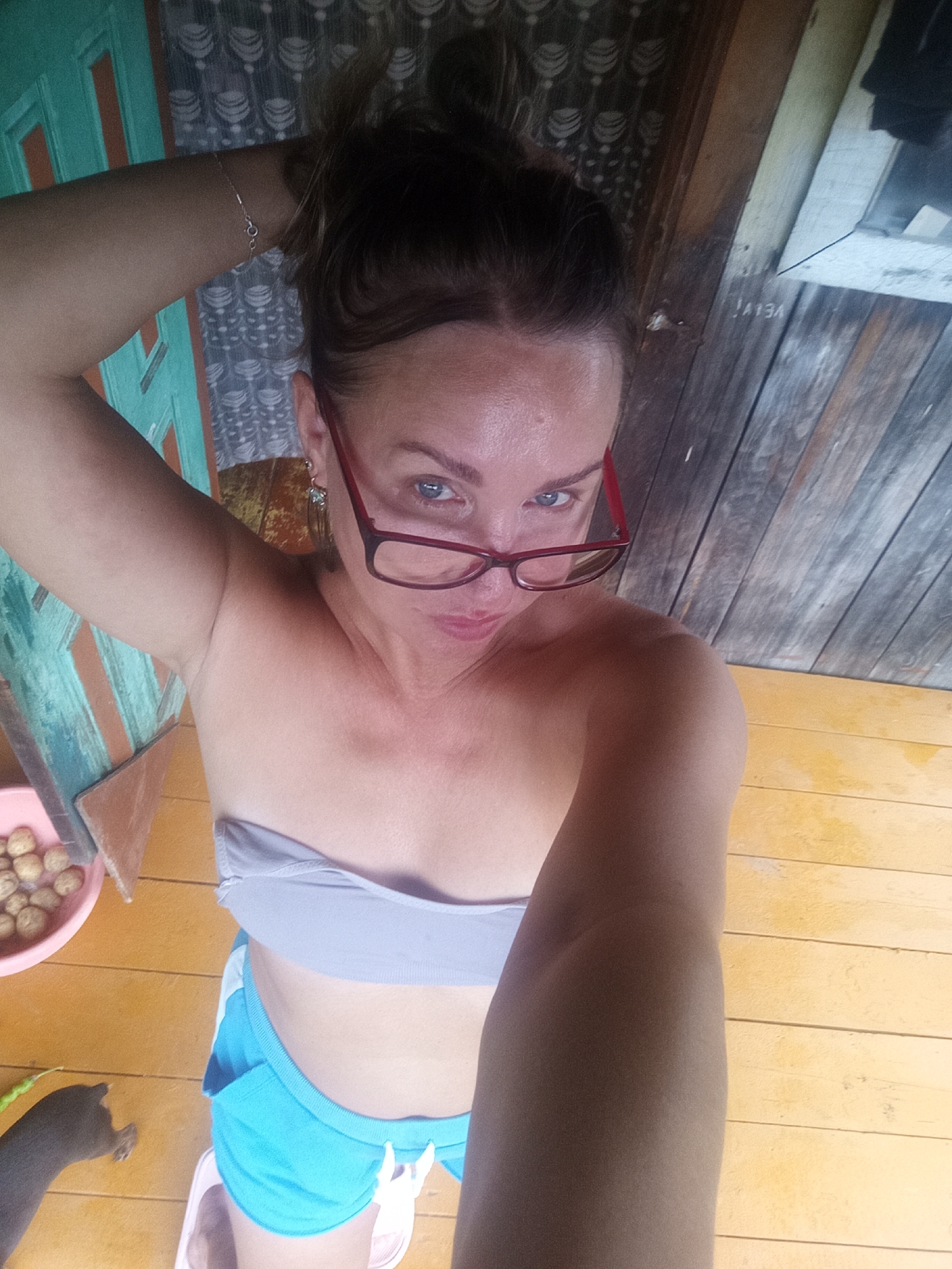 A woman is taking a selfie while lying down. She has brown hair pulled up in a bun, blue eyes, and is wearing red-rimmed glasses. She is wearing a light gray bandeau top and bright blue shorts. Her left arm is raised and resting on her head. Behind her, a wooden wall is visible with horizontal planks. To her left, a teal and orange striped panel and a pink bowl filled with round, light-colored objects are present. A small green object, possibly a bean or pea pod, is on the yellow wooden floor near the bowl. A dark-colored object, possibly a dog's paw, is visible in the lower left corner of the image. The floor is made of yellow wooden planks. The image does not provide any information to determine its location in the city of Novopokrovka, Russia.
