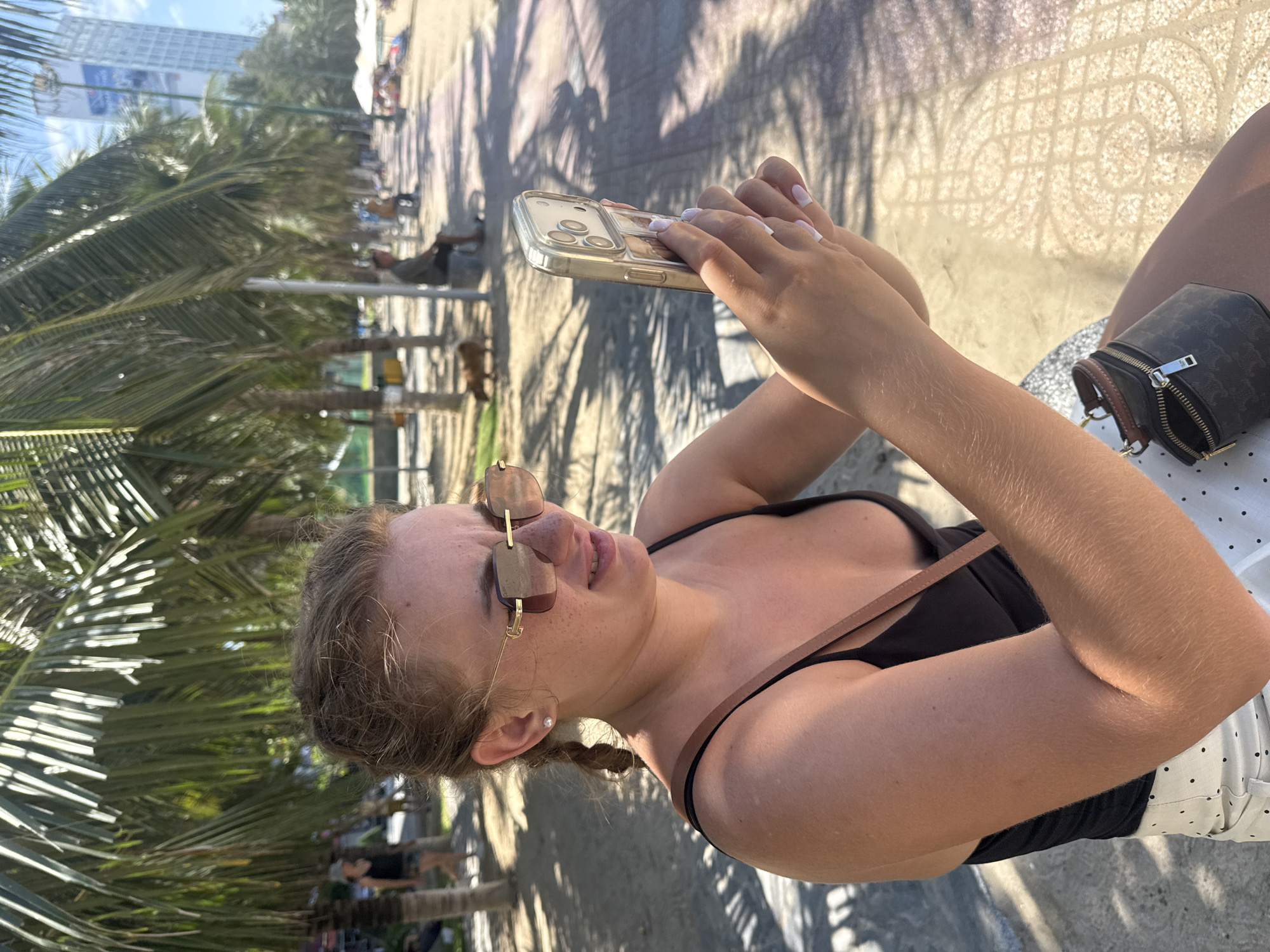In Nha Trang, Vietnam, a young woman with blonde hair pulled into a braid and wearing a black tank top and white polka-dot skirt sits on a concrete bench, holding a smartphone in her hands. She is wearing large, square-framed sunglasses and a small pearl earring. The woman appears to be looking at the phone, with her fingers positioned on the screen. Beside her, on the bench, is a small dark brown cross-body bag with a patterned design and gold hardware.
The background suggests a public area, likely a promenade or sidewalk adjacent to a beach. Palm trees with their fronds casting shadows dominate the upper portion of the image. Further in the background, other people are visible walking or sitting, and there are some beach umbrellas and a tall building can be seen in the distance. The ground is paved with tiles, and there is sand visible. The lighting indicates it is daytime, with the sun casting distinct shadows.