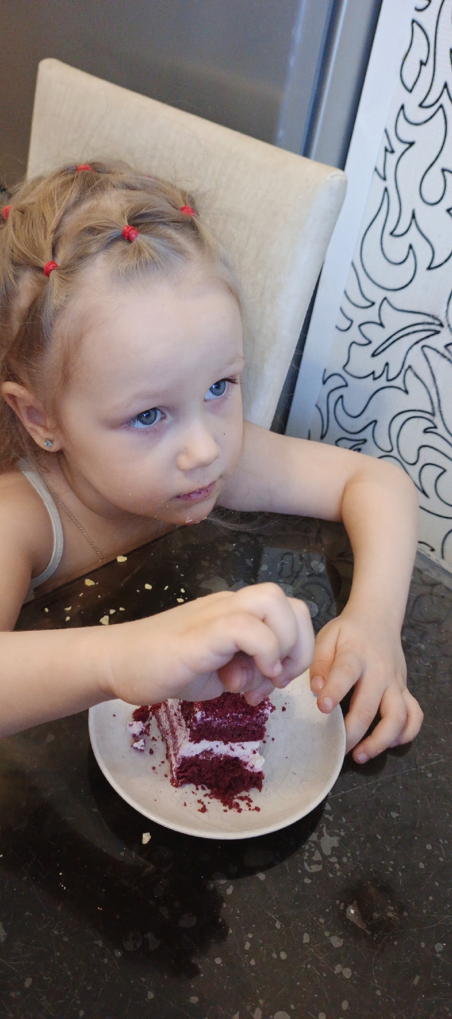 An indoor scene in Chaikovsky, Russia, depicts a young child seated at a dark, mottled countertop. The child has fair skin, blue eyes, and light blonde hair styled into multiple small pigtails secured with red elastic bands. A small star-shaped earring is visible in the child's left ear. The child wears a light-colored tank top and a thin chain necklace. Cake crumbs are present around the child's mouth and chin. The child's right hand is positioned over a light-colored plate, actively picking at a piece of red velvet cake with white cream frosting. Cake crumbs are scattered on the dark countertop around the plate. The child's left forearm rests on the table, and the child is looking upward and to the right. Behind the child, a light-colored, upholstered chair back is visible. To the right of the child, a white wall or panel with black, flame-like or abstract decorative patterns is present. The background to the left is a dark, indistinct surface.