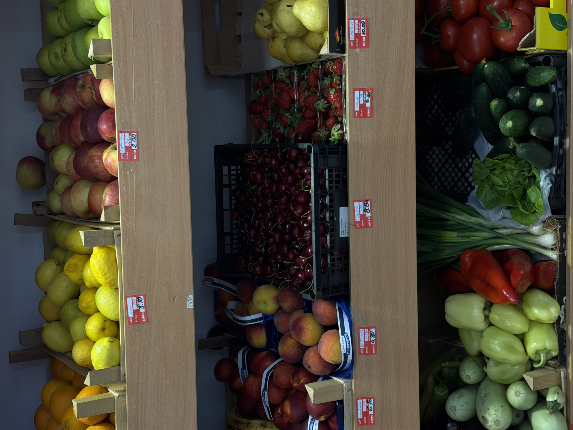 The image displays a supermarket produce aisle with shelves stocked with various fruits and vegetables. On the top shelf, there are crates of lemons and oranges. The middle shelf holds apples, peaches, and cherries. The bottom shelf contains zucchini, bell peppers, green onions, lettuce, cucumbers, and tomatoes. Price tags are visible in front of some of the produce. The scene appears to be a standard retail environment. Based on the price tags which show prices in "EURO" and the presence of Cyrillic script on some labels, this market is likely located in Budva, Montenegro, a popular tourist destination where Euro is widely used and Cyrillic is a common script.