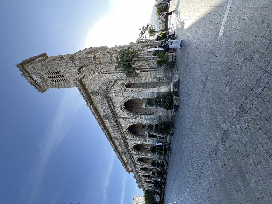 An exterior, low-angle, wide-shot photograph captures the stone facade of Nha Trang Cathedral (Christ the King Cathedral) in Nha Trang, Vietnam, under a clear blue sky. The Neo-Gothic style church, constructed from large grey stone blocks, dominates the frame, extending diagonally from the left foreground to the right background. Its prominent square bell tower, featuring a visible clock face displaying approximately 10:45, rises on the right side.
The church's long side elevation, visible on the left, features a series of ornate pointed arches supported by robust columns. White statues are visible within some of these arched recesses. The base of the building is lined with several potted evergreen trees, trimmed into conical shapes. Further along the left, several motorcycles are parked alongside the building. The ground in the foreground and extending towards the church is a wide plaza paved with grey rectangular stone blocks.
On the right side of the image, bright, direct sunlight creates strong illumination and casts distinct shadows across the paved plaza. Multiple palm trees are visible near the church entrance and in the background. Several individuals are present: one person in a dark top and light pants stands in the mid-ground on the right, partially illuminated by the sun. Other smaller figures are discernible in the far background on a higher elevation, suggesting a hillside or terrace. A tall residential building is partially visible in the distant background on the far left.