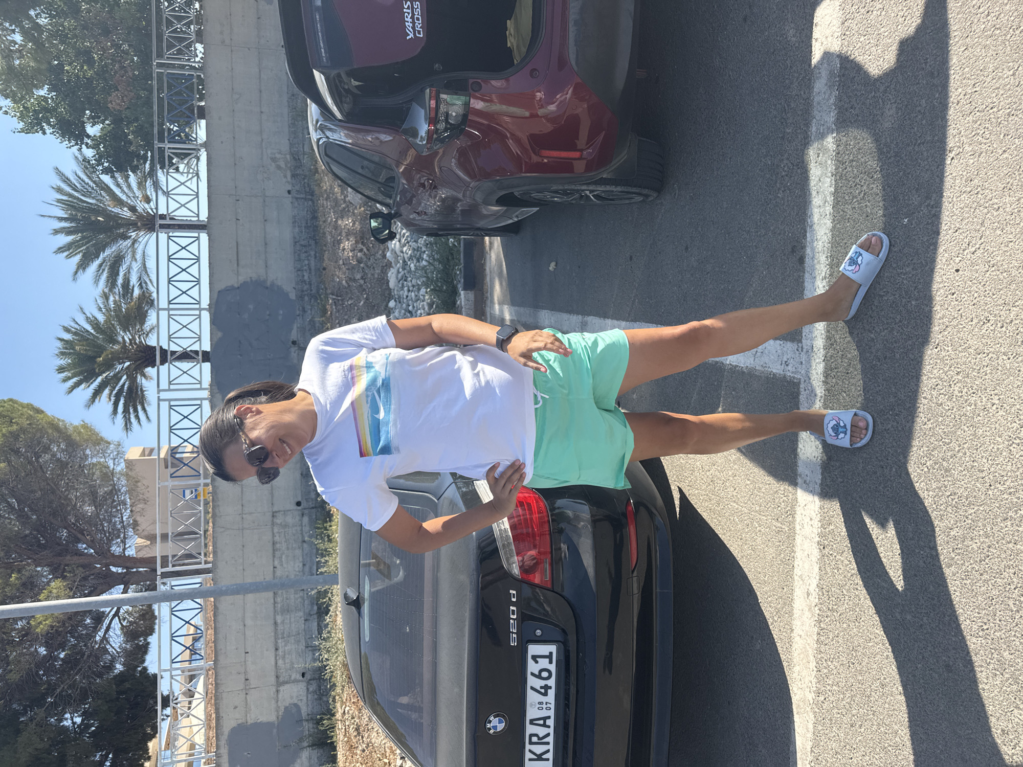 A woman stands in a parking lot next to two cars, a black BMW 520d and a dark red Toyota Yaris Cross. The woman is wearing a white t-shirt with a rainbow-colored graphic, light green shorts, and light blue slides with a cartoon character. She has dark hair pulled back and is wearing sunglasses. The background features a concrete wall, a railing with a geometric pattern, palm trees, and other vegetation, under a clear blue sky. The license plate of the BMW is visible and reads "KRA 461". The scene suggests a location in Polis Chrysochous, Cyprus.