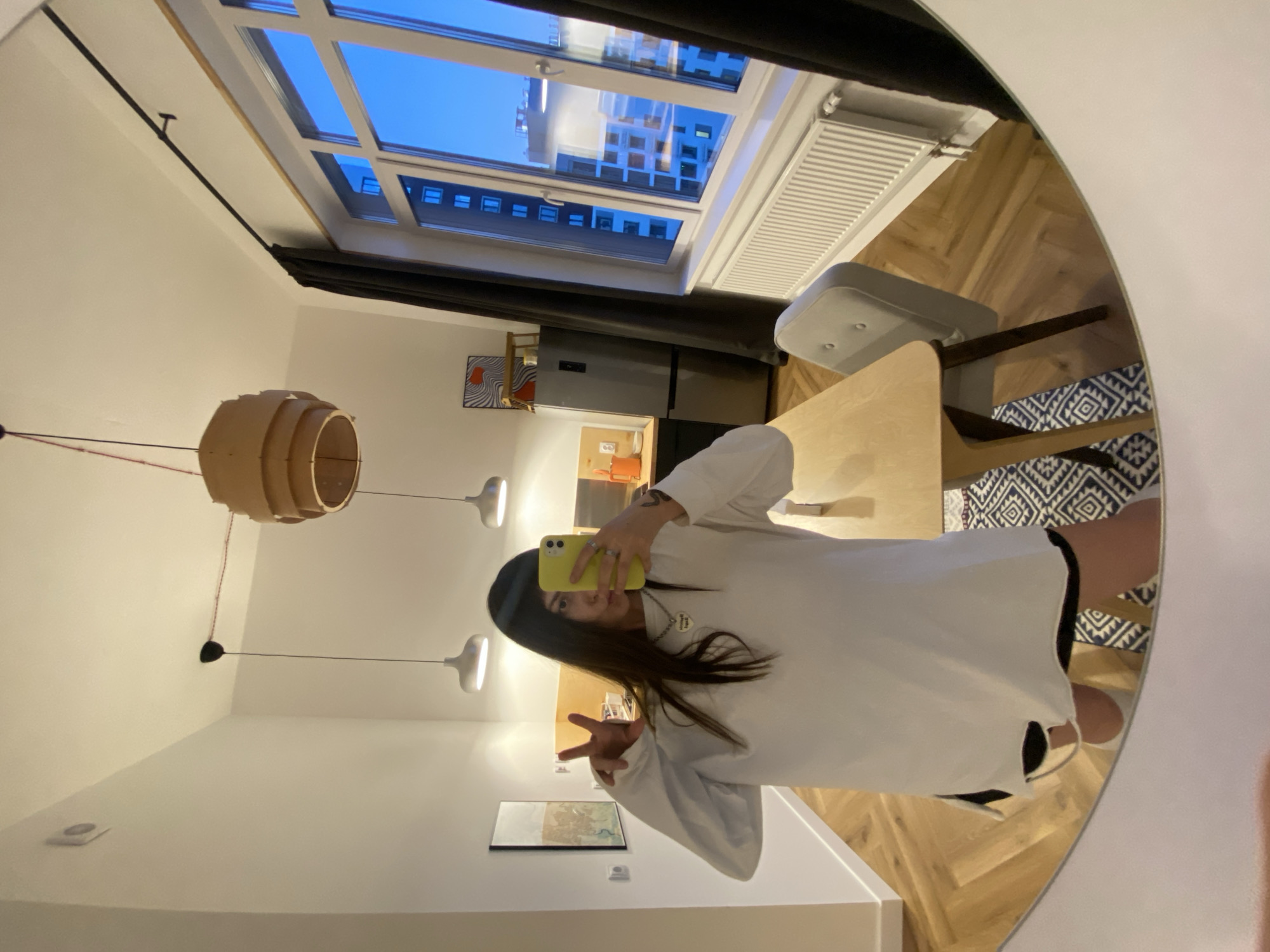 A person is taking a selfie in a mirror inside a modern apartment. The person is wearing a white oversized sweatshirt and a black beanie and is holding a yellow smartphone up to their face, using it as a mirror to take the picture. The person is making a peace sign with their left hand.
The apartment appears to have a kitchen and dining area. Visible features include:
* A table and chairs.
* A modern light fixture hanging from the ceiling.
* A window that reveals a view of an urban environment outside.
The city outside appears to be Izhevsk, Russia. The buildings visible through the window seem consistent with the architecture found in parts of Izhevsk.
Other details include a white radiator, a painting of a person, a fridge, and a patterned rug in the apartment. The apartment's design suggests a modern and minimalist style. The time of day is likely evening or night, as evidenced by the outside light.