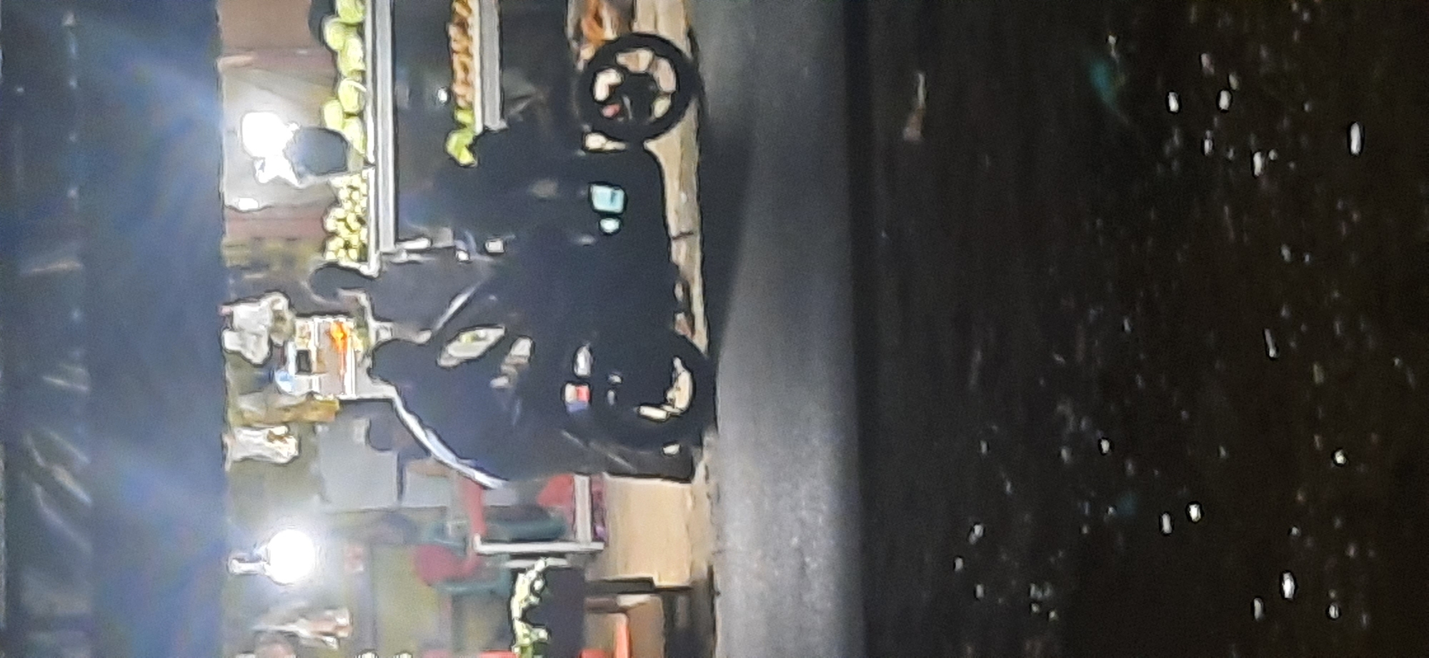 The image depicts a dimly lit street scene, likely in Cisauk, Indonesia, with a focus on a motorcycle and two individuals. The scene appears to be in the evening or at night, with artificial lighting from a nearby stall or building illuminating the area. In the foreground, a dark-colored motorcycle is parked. A person, silhouetted and leaning over the motorcycle, is visible. Another person stands behind the motorcycle. Behind them, a stall displays various items, possibly fruits or vegetables, under bright lighting. The ground is paved, and there are reflections of lights on the wet pavement in the lower portion of the image. The overall impression is that of a roadside vendor or a gathering at night.
