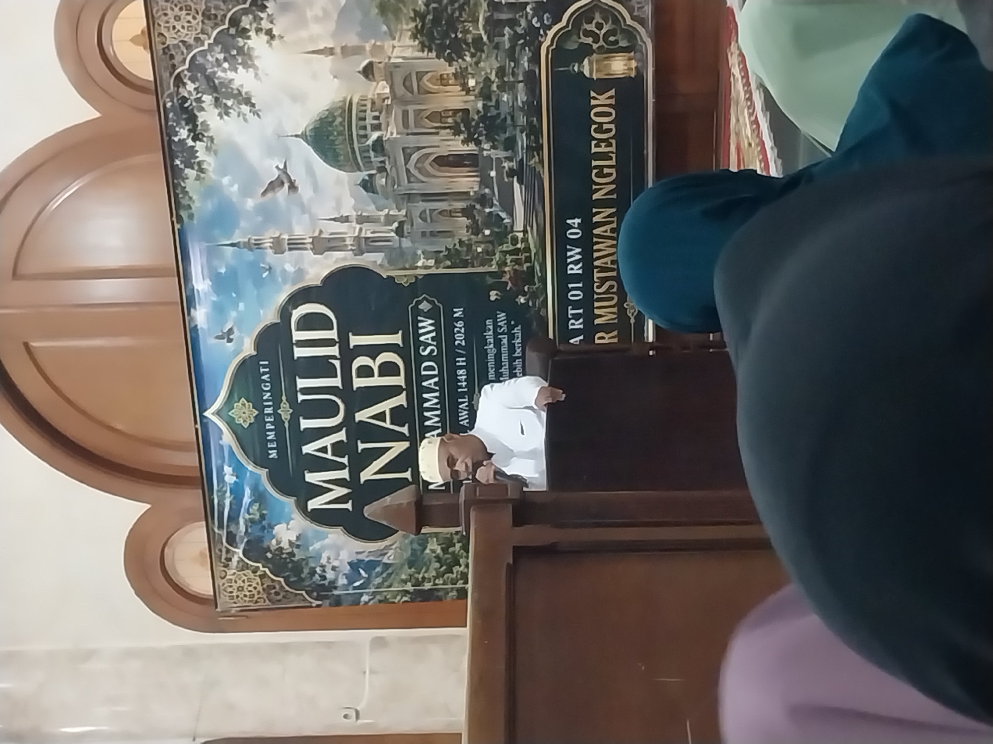 In an indoor setting, likely a mosque or community hall in Nglegok, Indonesia, a man wearing a white cap and white attire is speaking into a microphone while standing behind a wooden podium. He is positioned in front of a large banner displaying "MAULID NABI MUHAMMAD SAW" and an illustration of a mosque and minarets. Several people, mostly women wearing headscarves, are seated in the foreground, facing away from the camera, suggesting they are attending an event. The banner also contains text that appears to be in Indonesian, possibly an address or event details, including "RT 01 RW 04" and "R MUSTAWAN NGLEGOK." The ambiance suggests a religious gathering or celebration.