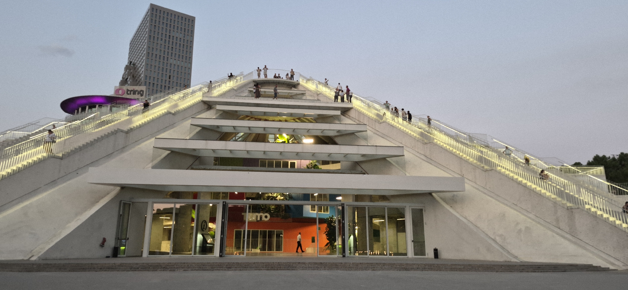 The image shows the exterior of the Tring building in Tirana, Albania, with its distinctive tiered, pyramid-like structure leading up to the main building. A large, modern skyscraper is visible in the background to the left of the Tring building. The Tring building itself features wide, illuminated staircases on either side of the structure, with numerous people scattered across them, some walking, some standing, and some sitting. At the base of the structure, large glass doors reveal the colorful interior of what appears to be a commercial space, with signage that includes the word "Tring" in orange letters. The sky is overcast and the lighting suggests it is either early morning or late evening. The overall impression is of a public space designed for people to gather and ascend to a higher level.