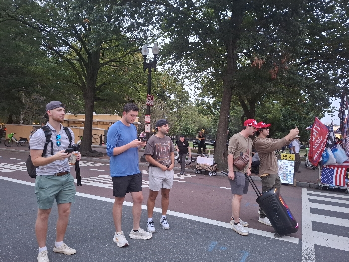 The image depicts an outdoor urban scene during daylight hours, consistent with a street bordering Lafayette Square Park in Washington, D.C., United States.
Six male individuals are prominently visible in the foreground and mid-ground. From left to right:
1. A male in a grey backward baseball cap, striped polo shirt, green shorts, and white sneakers stands on the asphalt, holding a black remote-like device and a white object. A black backpack is worn, and blue sunglasses hang from his shirt.
2. A male in a blue long-sleeve shirt, dark shorts, and white sneakers stands on the asphalt, looking down at a smartphone in his hands.
3. A male in a patterned t-shirt, light shorts, and a dark backward baseball cap, wearing white sneakers, stands on the asphalt, looking towards the right.
4. A male in a red "TRUMP" baseball cap, olive-green long-sleeve shirt, grey shorts, and light sneakers, with a brown sling bag, stands on the asphalt adjacent to a crosswalk.
5. A male in a red "TRUMP" baseball cap and a brown long-sleeve shirt stands near the crosswalk, extending both arms forward to hold a smartphone horizontally, appearing to record or take a selfie. He is simultaneously pulling a black, wheeled speaker unit featuring illuminated red, blue, and green lights on its side.
The environment includes an asphalt street with white crosswalk markings on the right and dashed white lines on the left. A reddish-brown paved area serves as a sidewalk or plaza. Numerous mature trees with green foliage form a dense canopy. A multi-lamp street light is visible. In the background, a light-colored building with tan walls is partially visible, along with parked green and white shared bicycles. On the far right, near the crosswalk, a vendor stall displays multiple flags, including several red "TRUMP" flags (some stating "TRUMP 2020"), a red flag with white text, and an American flag. Other merchandise is also visible on the stall. Several indistinct figures and small wagons are discernible in the deeper background.