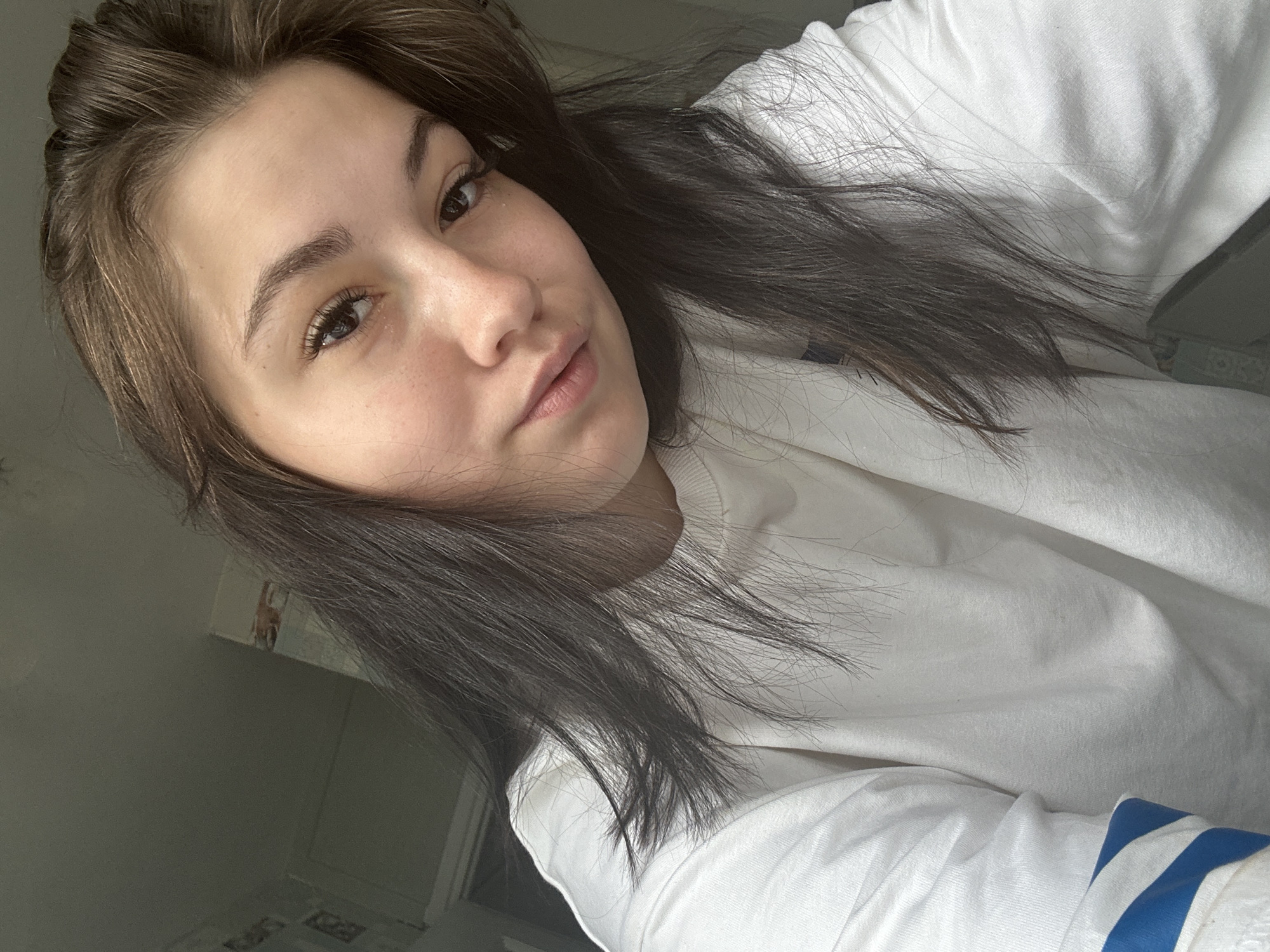 A young woman is captured in a selfie, her face angled slightly towards the viewer. She has long, dark brown hair that falls loosely around her shoulders. Her skin appears fair with subtle freckles. She is wearing a plain white crewneck sweatshirt with blue stripes on the sleeves, partially visible. The background is dimly lit and appears to be an indoor setting, possibly a room. The lighting suggests it may be daytime, with light coming from the left side of the frame, casting soft shadows. The image is framed from the chest up, focusing on her facial features and upper body. There is no discernible information indicating a specific location within the city of Chaikovsky, Russia, or any urban elements in the background.