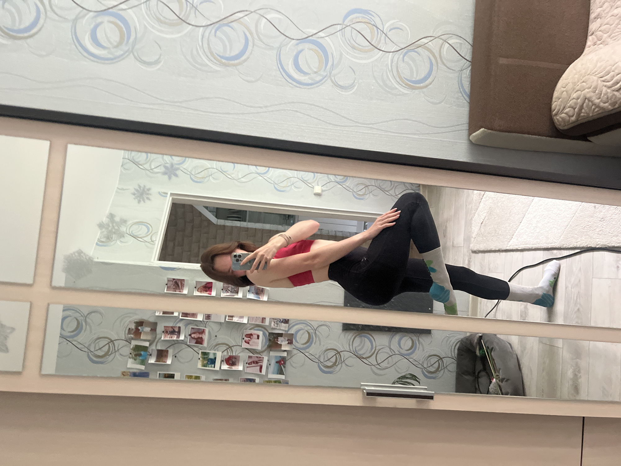 A woman is visible in a mirror selfie. She is wearing a red top and black leggings, with patterned socks on her feet. The background reveals a room with patterned wallpaper, photographs attached to the wall, a doorway, and a glimpse of furniture including a sofa. While the photo was taken in Chaikovsky, Russia, the exact location within the city cannot be determined from the image.