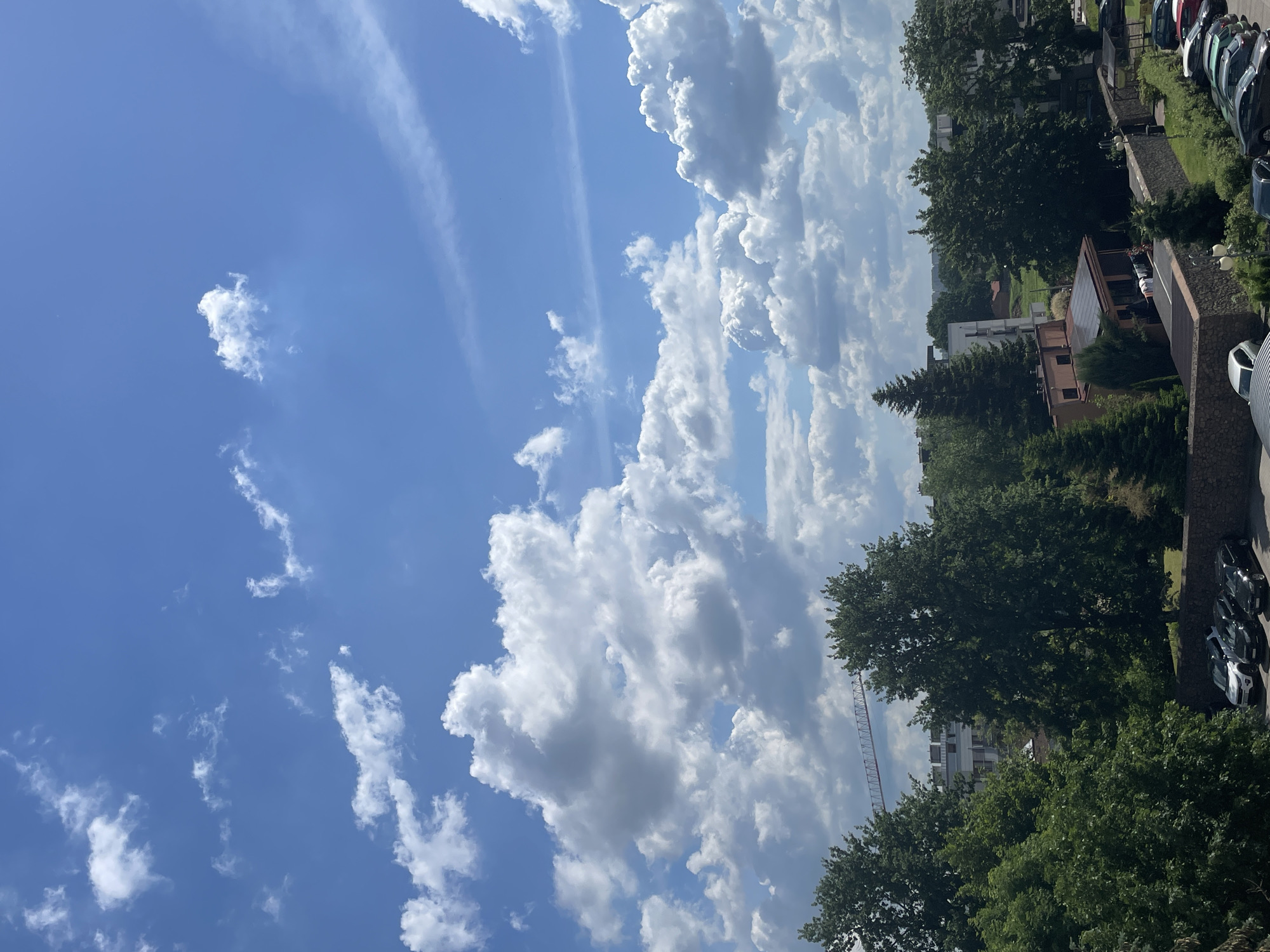 The image displays a bright, clear blue sky filled with various white clouds, some appearing fluffy and cumulus, others more wispy and cirrus-like. In the lower portion of the image, a cityscape is visible. Lush green trees with full canopies dominate the foreground and midground, interspersed with buildings. A construction crane is prominent against the sky in the distance, suggesting ongoing development. Below the trees and buildings, a parking lot is visible with several cars parked, including SUVs and sedans in black, white, and silver. The scene is framed by a stone wall in the lower right, with some parked cars visible beyond it. The overall impression is a sunny day in a developed area with abundant greenery.