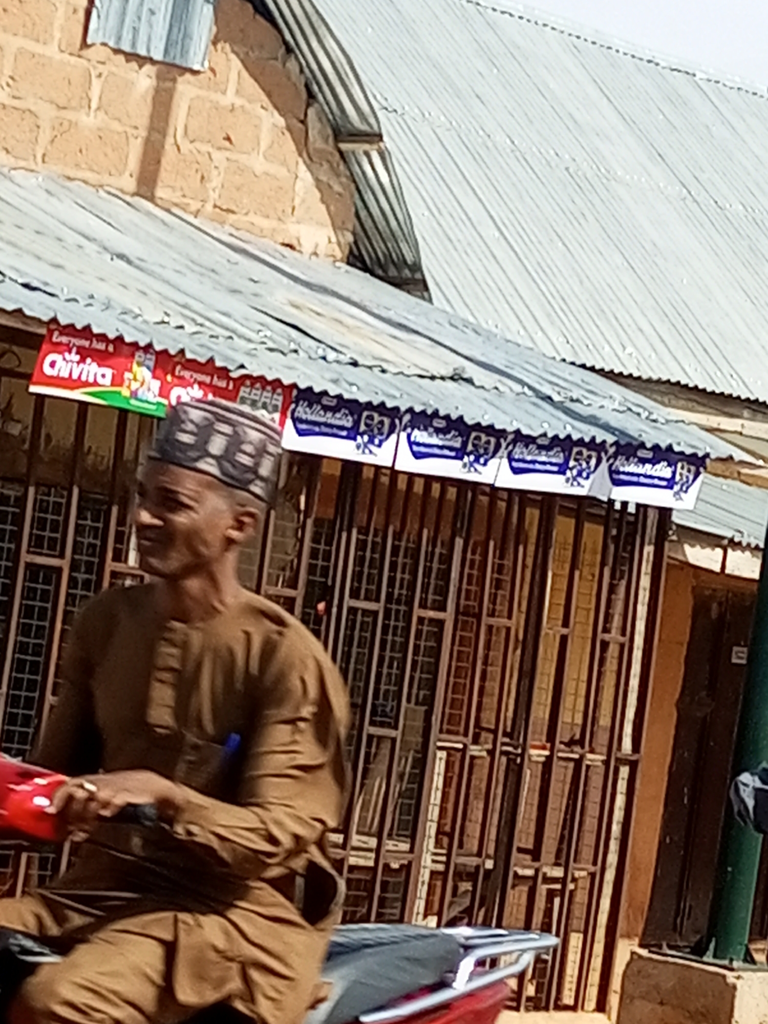The image shows a man wearing a brown tunic and a patterned cap, sitting on what appears to be a motorcycle. He is looking to his left, with a faint smile. In the background, there is a building with a corrugated metal roof and brick walls. Signs for "Chivita" and "Hollandia" are visible above the building's windows. The windows themselves are barred, suggesting a secure or older structure. The scene appears to be outdoors, likely in a street or marketplace in Tempure, Nigeria, given the architecture and signage commonly found in such locations. The lighting suggests it is daytime.