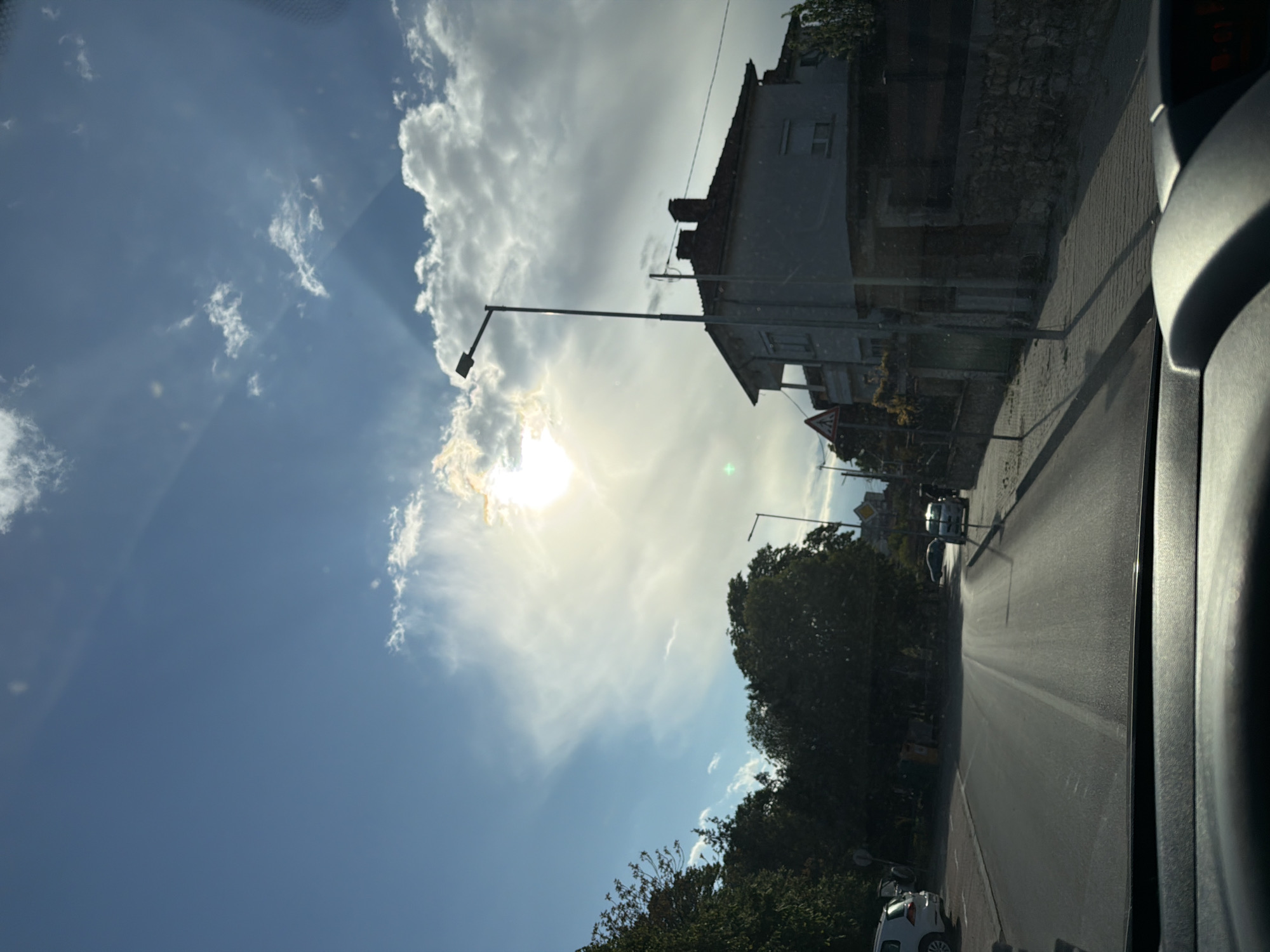 The image is a tilted roadside view of a street, taken from inside a vehicle. The sky is partially covered with clouds, and the sun shines through them. A building stands beside the street. Streetlights line the road. Cars travel along the street. Trees grow near the roadside.