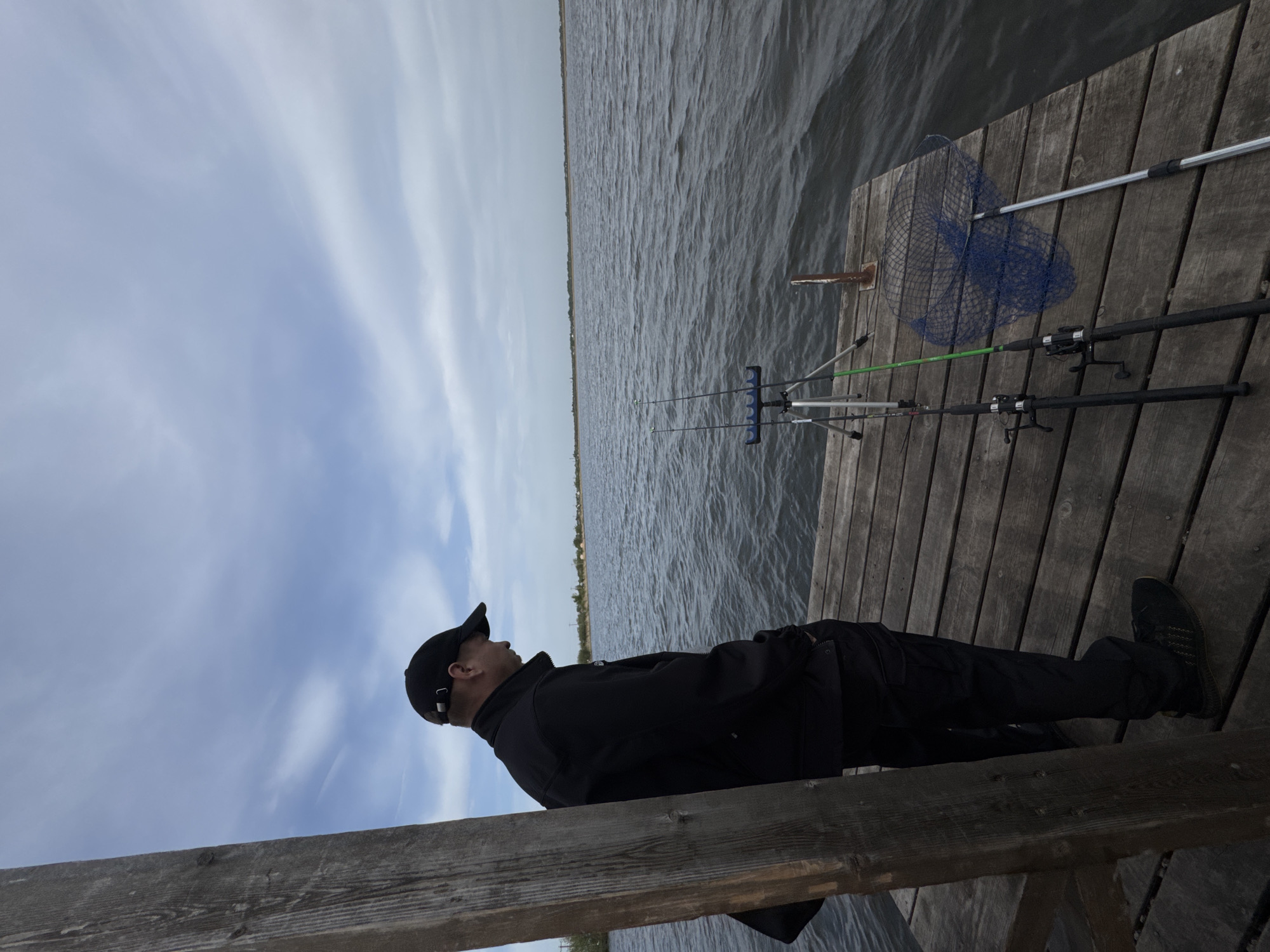 A man is lying on a wooden pier next to a body of water under a cloudy sky. The man is wearing a black jacket, black pants, a black baseball cap, and black shoes. Fishing rods and a fishing net are lying on the pier near him. The location is Unknown, Unknown.