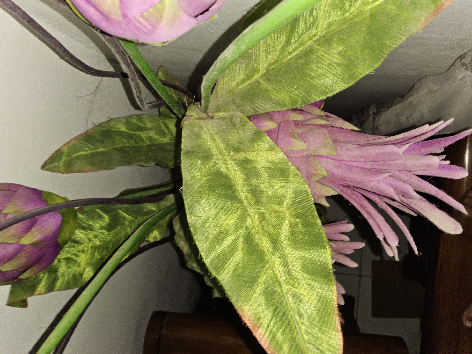 The image is a close-up shot of artificial pink flowers and green leaves against a light-colored wall. The flowers have a drooping, bell-like shape with delicate, fuzzy petals. The leaves are broad and veined, with a slightly textured surface. The overall lighting is dim, casting shadows and highlighting the textures of the artificial plants. There are no people or animals visible in the image. The background suggests an indoor setting, possibly a home or a decoration in a public space. There is no visible indication of the image being taken in Payagaboh, Indonesia.
