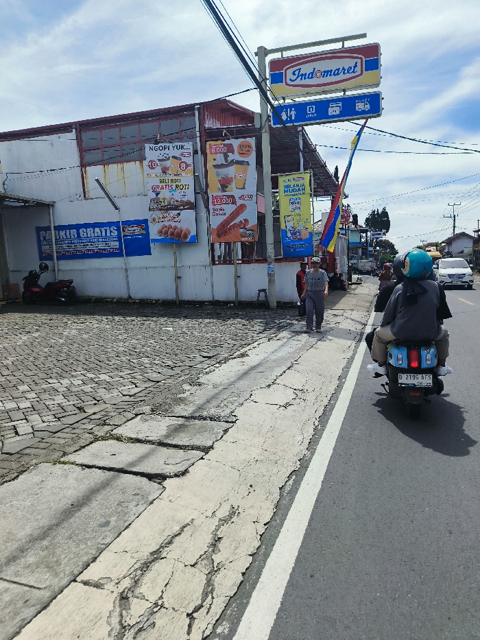 The scene is taken in Ciwidey, Indonesia at a location with coordinates -7.108 latitude and 107.454 longitude. A blue motorcycle is parked on the right side of the asphalt road, facing the camera. The motorcycle has a license plate that reads “D 2136 DFS.” Two people are seated on the motorcycle. One person is wearing a blue helmet and a dark jacket. The other person is wearing a black jacket and a white helmet. Behind the motorcycle, on the right side of the road, there are two other people. The person in the front is wearing a black jacket and a white helmet. The person behind is wearing a black jacket and a black helmet. In the background, there is a building with a red roof and a white sign. The sign reads “Indomaret” in red and white letters. The building is also adorned with advertisements for various products, including hot dog buns, and drinks. There is a cobblestone area to the left of the image, which may be a parking lot. The sign reads “Parkir Gratis,” which means “Free Parking.” The sky is partly cloudy, with a bright blue sky and a few white clouds.