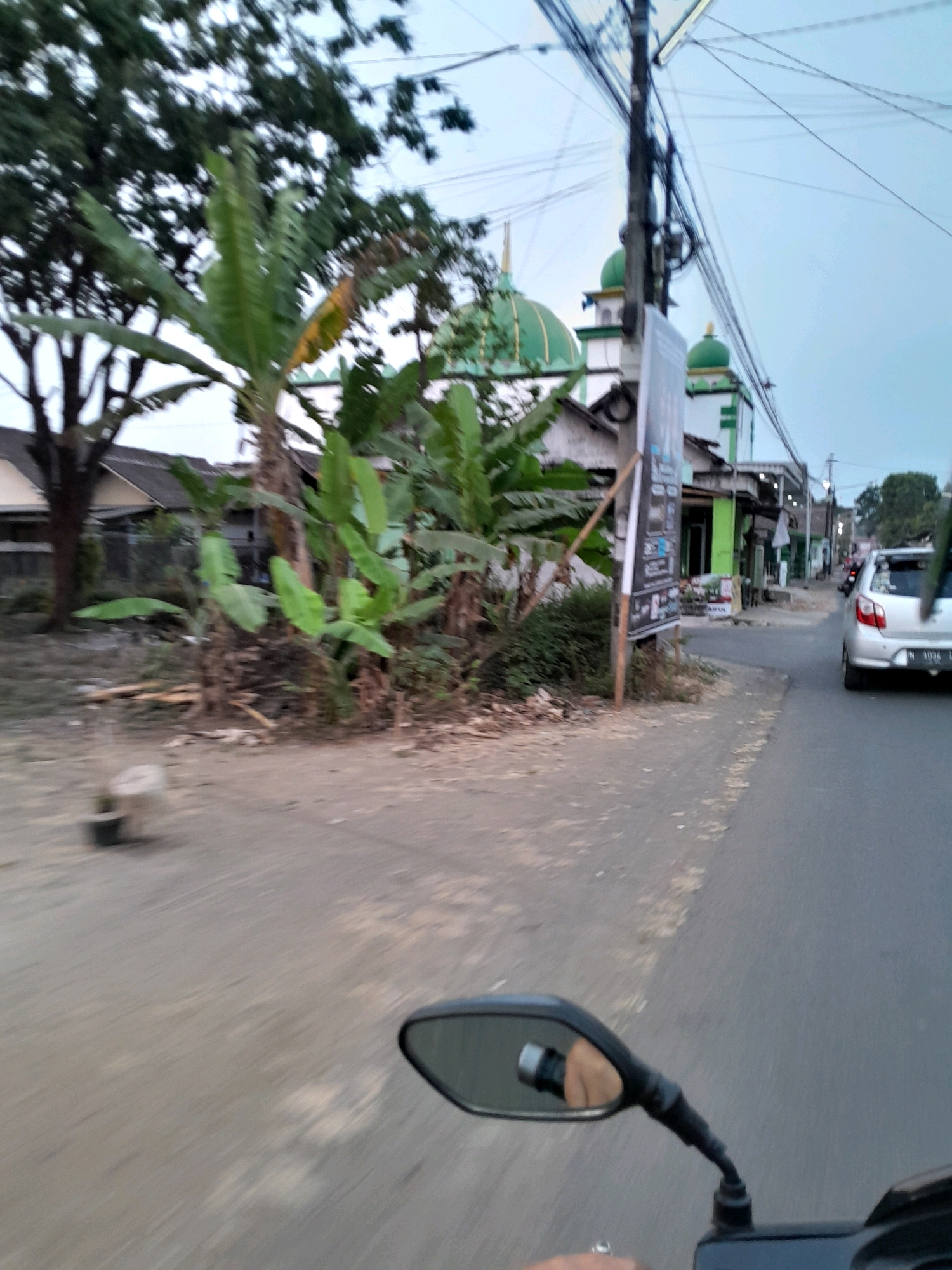 In Dilem, Indonesia, a street view captures a paved road with a silver car visible on the right. Adjacent to the road is a dirt path, and further back, trees and buildings are present. Two green-domed mosques with white structures are prominent in the background. A utility pole with numerous wires stands near the mosques. The scene appears to be captured from a moving vehicle, likely a motorcycle, as indicated by the presence of a motorcycle's rearview mirror in the foreground.