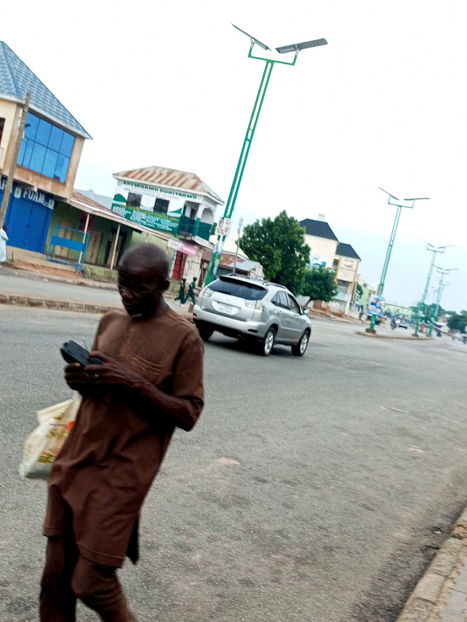 An outdoor, eye-level, medium shot captures an elderly man in a brown dashiki and pants, walking on a paved street in Gombe, Nigeria. He holds a black mobile device in his hands and a white bag with red and green accents is slung over his left shoulder. The man is in the foreground, slightly blurred, suggesting movement. In the background, a silver SUV is parked on the right side of the street, facing away from the viewer. To the left, a two-story building with a blue-tinted glass facade stands beside a smaller building with a corrugated metal roof. The street is lined with modern, green solar-powered streetlights. The sky is overcast and bright. The overall scene depicts an ordinary day on a street in Gombe, a city in northeastern Nigeria.