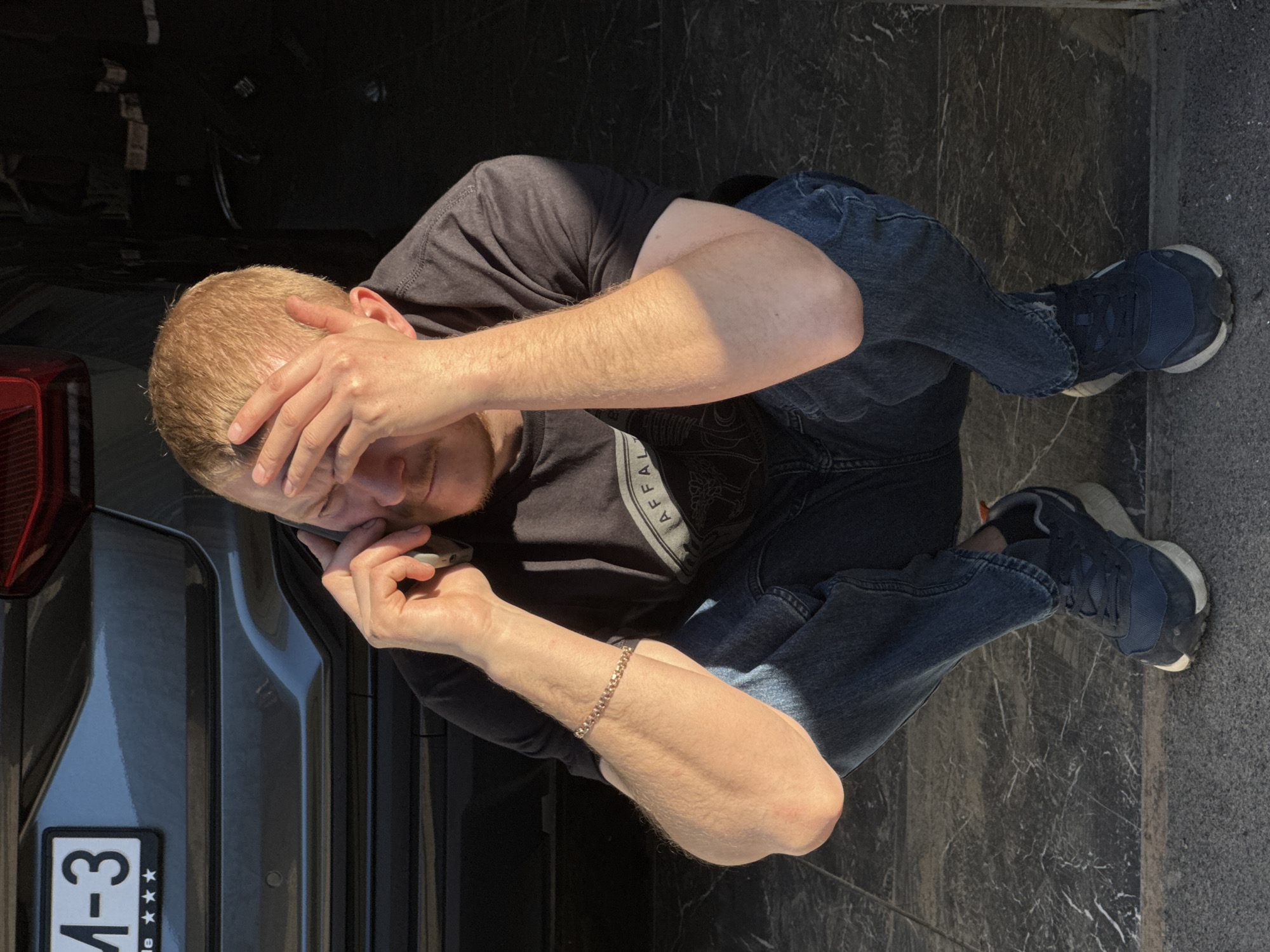 A man is crouched on a gray marble-tiled surface next to a dark-colored vehicle, apparently in Chaikovsky, Russia, judging from the license plate that begins with "M-3". He is wearing blue jeans, a black t-shirt, and dark blue athletic shoes. He is holding a mobile phone up to his ear with his right hand while his left hand is placed on his head. The lighting suggests an outdoor location with strong sunlight casting shadows. The vehicle is positioned to his left, and the marble tiling extends behind and to his right.