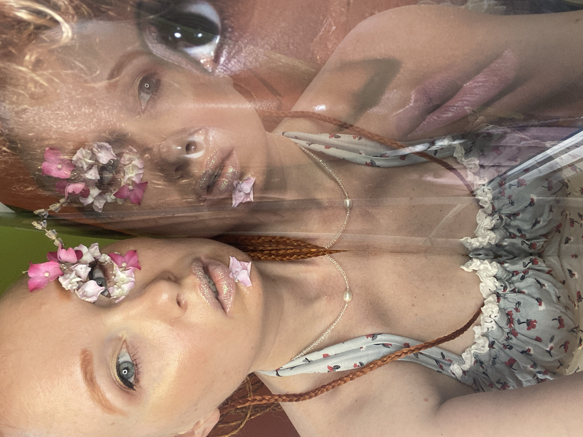This is a close-up, double-exposed portrait of a fair-skinned woman with reddish-blonde hair and braided pigtails. She is wearing a light blue floral print top with white lace trim. There are small pink and white flowers near her left eye and on her lower lip. Her lips are glossy and appear to have glitter applied. She is wearing a delicate necklace with a pearl pendant. The background is a blurred green and orange. The image is a creative portrait with the woman's face appearing in two slightly offset layers, creating a ghostly effect. There is no visible location information indicating Sochi, Russia.