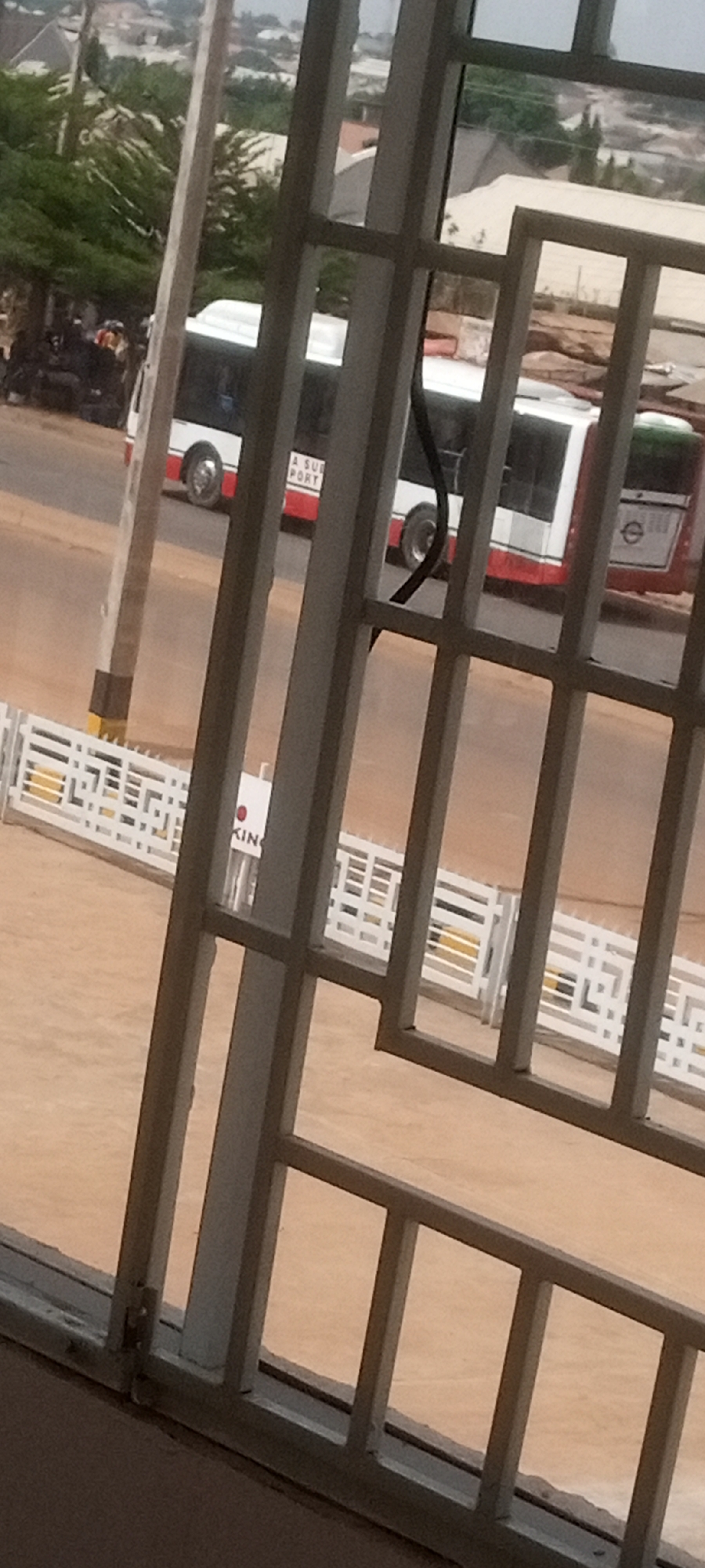 A partial view through a gray metal-framed window reveals an urban street scene in Sabon Tasha, Nigeria. A white bus with red trim is visible through the window bars, partially obscured. A group of people is gathered to the left of the bus. A white decorative fence with a yellow base runs parallel to the window and a white sign with "KING" on it is next to the fence. The sky is overcast and other buildings are visible in the distance.