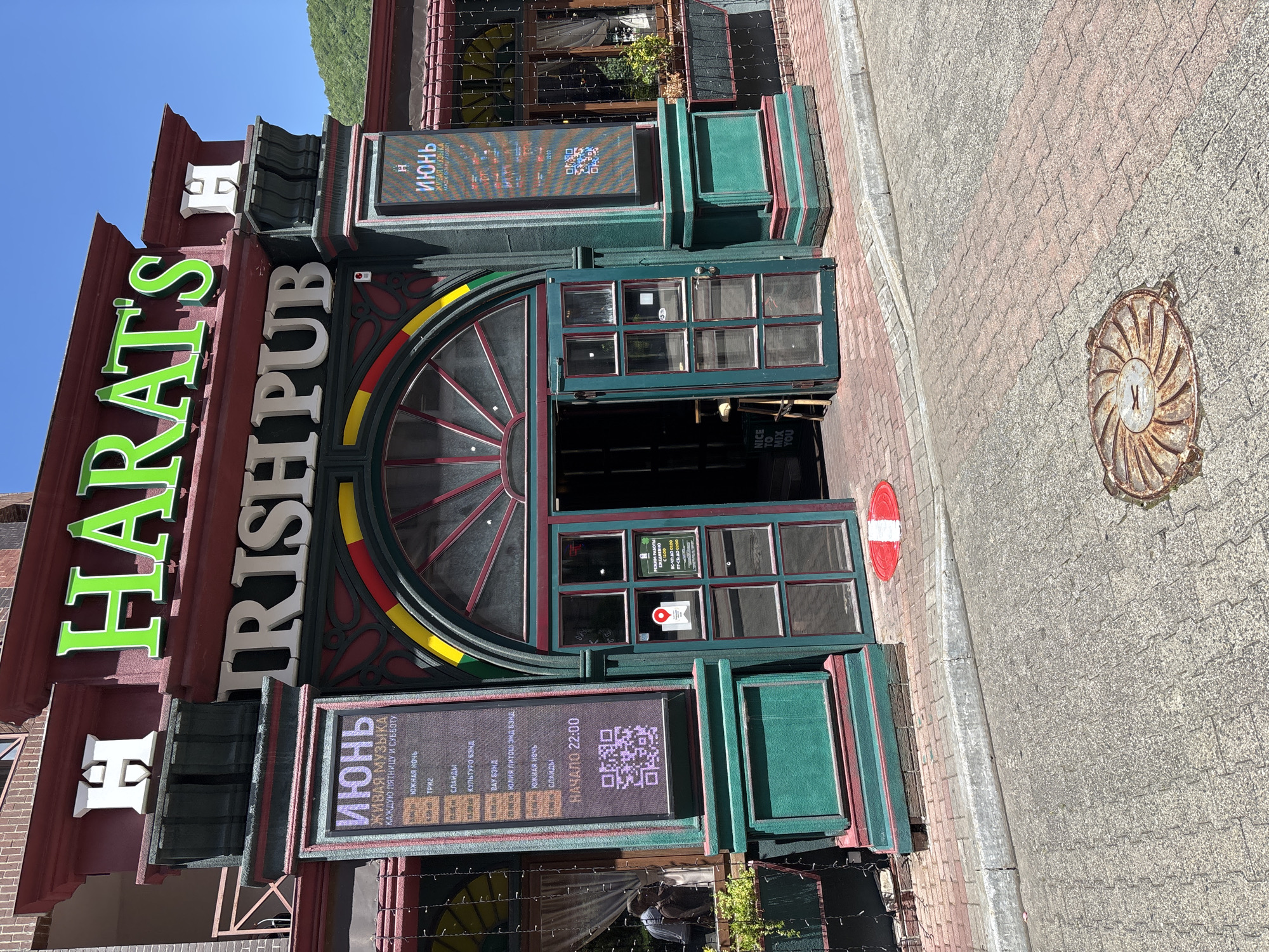 The image shows the exterior of "Harat's Irish Pub" located in Estosadok, Russia. The pub has a prominent green sign with "HARAT'S" in large, stylized letters. Below this, "IRISH PUB" is written in white. The entrance features a large arched window with stained glass elements in red, yellow, and green. Double doors, also with glass panes, are open, revealing a dark interior with a stool.
To the left of the entrance, a digital display board shows an advertisement in Cyrillic for "ИЮНЬ" (June) and "ЖИВАЯ МУЗЫКА" (Live Music), detailing various events and a start time of 22:00, along with a QR code. A smaller sign on the door reads "NICE TO MIX YOU". To the right of the entrance, another digital display board is visible, also showing advertisements.
The building's facade is painted in dark red and green, with decorative architectural elements. The ground in front of the pub is paved with reddish-brown bricks, leading to a grey concrete curb and a grey street. A rusty, circular manhole cover is present on the street. In the background, lush green hills are visible under a clear blue sky. The overall scene depicts a sunny day outside a lively establishment.