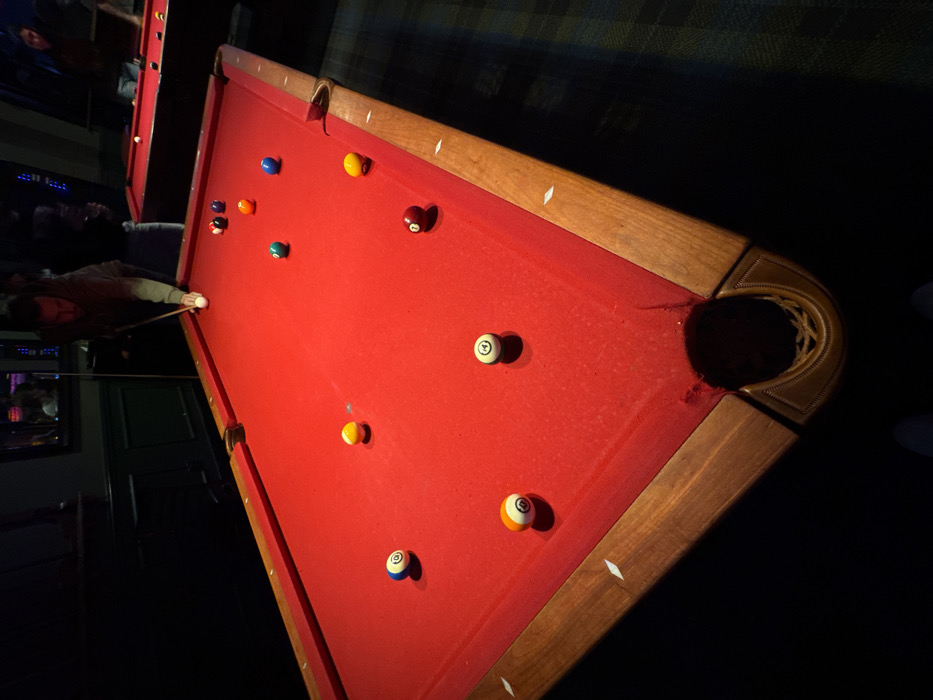 The image depicts an indoor scene within a dimly lit commercial establishment, likely a pool hall or bar, located in Los Angeles, United States of America. The specific address or neighborhood within Los Angeles is not discernible from the image.
The primary subject is a regulation-size pool table with worn red felt and dark wooden rails. Diamond-shaped markers are inlaid into the rails. A dark leather-like corner pocket is visible in the bottom right foreground. Scattered across the table are eleven billiard balls: a white cue ball positioned near the center, and ten numbered balls, including 1 (yellow/red), 2 (blue/white), 4 (purple/white), 5 (orange/white), 6 (green/white), 7 (dark red/white), 8 (black), 9 (yellow/white), and 10 (blue/white).
A male individual, appearing to be in his 20s or 30s with short dark hair and a beard, is leaning over the far side of the foreground pool table. He is wearing a dark, long-sleeved top and holding a pool cue, positioning it to strike the cue ball, indicating an active game.
In the background, another red-felted pool table is partially visible, also under localized lighting. At least two other individuals are faintly discernible near this background table, suggesting other games or social activity. The walls are dark, possibly paneled, and reflections from overhead lighting are visible in some areas. The floor is covered with a dark, patterned carpet. The overall lighting is low, with illumination concentrated directly above the pool tables.
