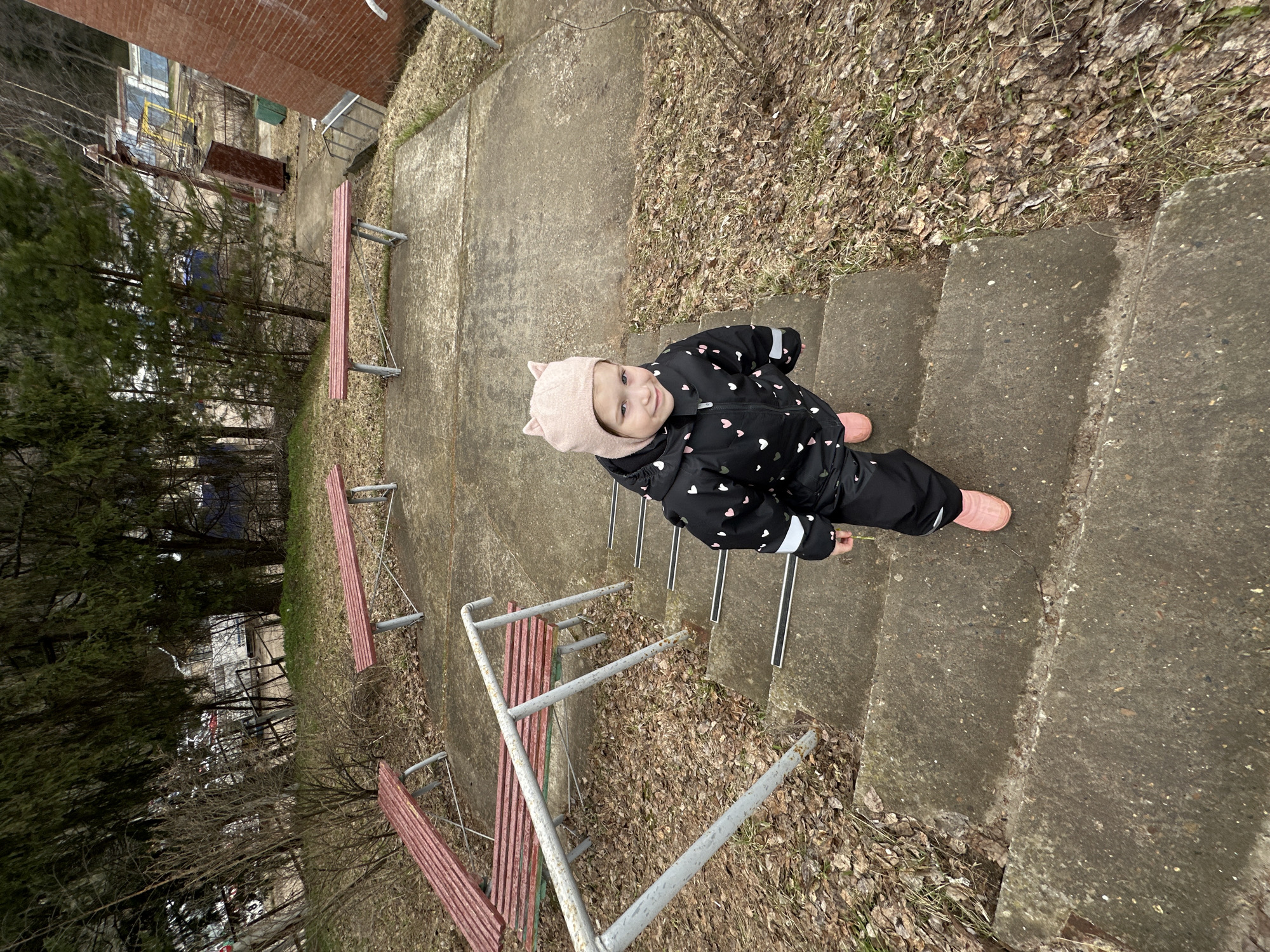 A young child in a black snowsuit with pink hearts and a pink hat with cat ears stands on concrete steps. The child is looking up and smiling. The setting appears to be a park or outdoor area with benches and trees. The location is Chaikovsky, Russia.