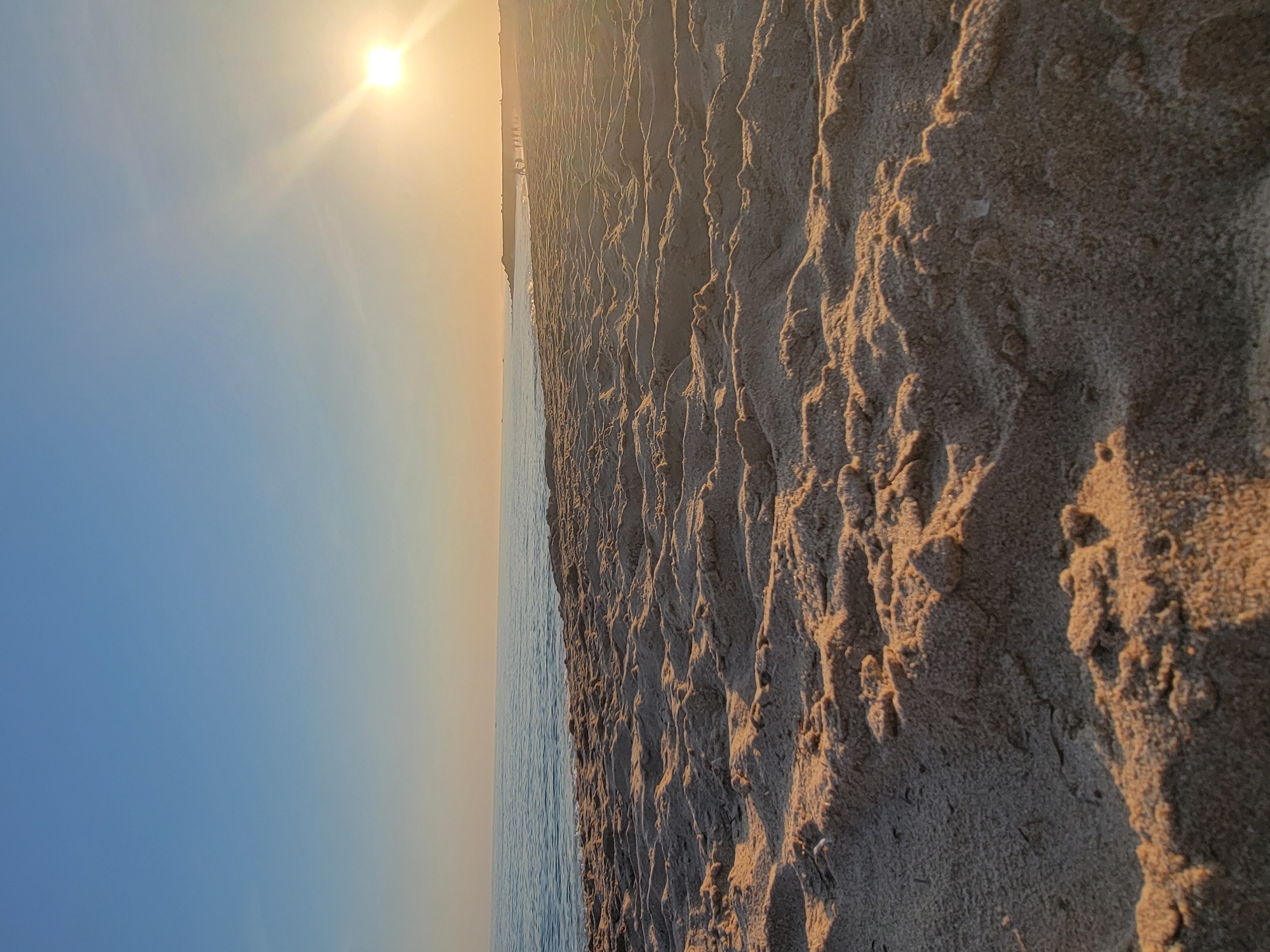 This media file captures a serene beach scene at sunset in Quarteira, Portugal. The image is oriented vertically, with the sun shining brightly in the upper left quadrant, casting a warm, golden glow across the sky and the sandy shore. The sun's rays are visible, creating a lens flare effect that adds to the ethereal atmosphere.
The foreground is dominated by the textured surface of the sand, showing intricate patterns created by the receding tide or wind. The sand is a light brown and tan color, with shadows accentuating its ruggedness. In the mid-ground, a calm body of water, likely the Atlantic Ocean, meets the shore, its surface reflecting the soft light of the setting sun. The water transitions from a gentle blue near the horizon to a darker hue closer to the beach.
In the far distance, a faint silhouette of a coastline is visible, possibly indicating buildings or a distant town, but these details are blurred and indistinct. The sky is a gradient of colors, starting with a clear, soft blue on the left and gradually transitioning to a pale orange and yellow near the horizon, a hallmark of a sunset.
No people or specific objects are clearly discernible in the image, emphasizing the natural beauty of the landscape. The overall mood is tranquil and peaceful, evoking a sense of calm and relaxation associated with a beach at dusk. The time of day is clearly sunset. The weather appears to be clear and calm. There is no visible text in the media file.