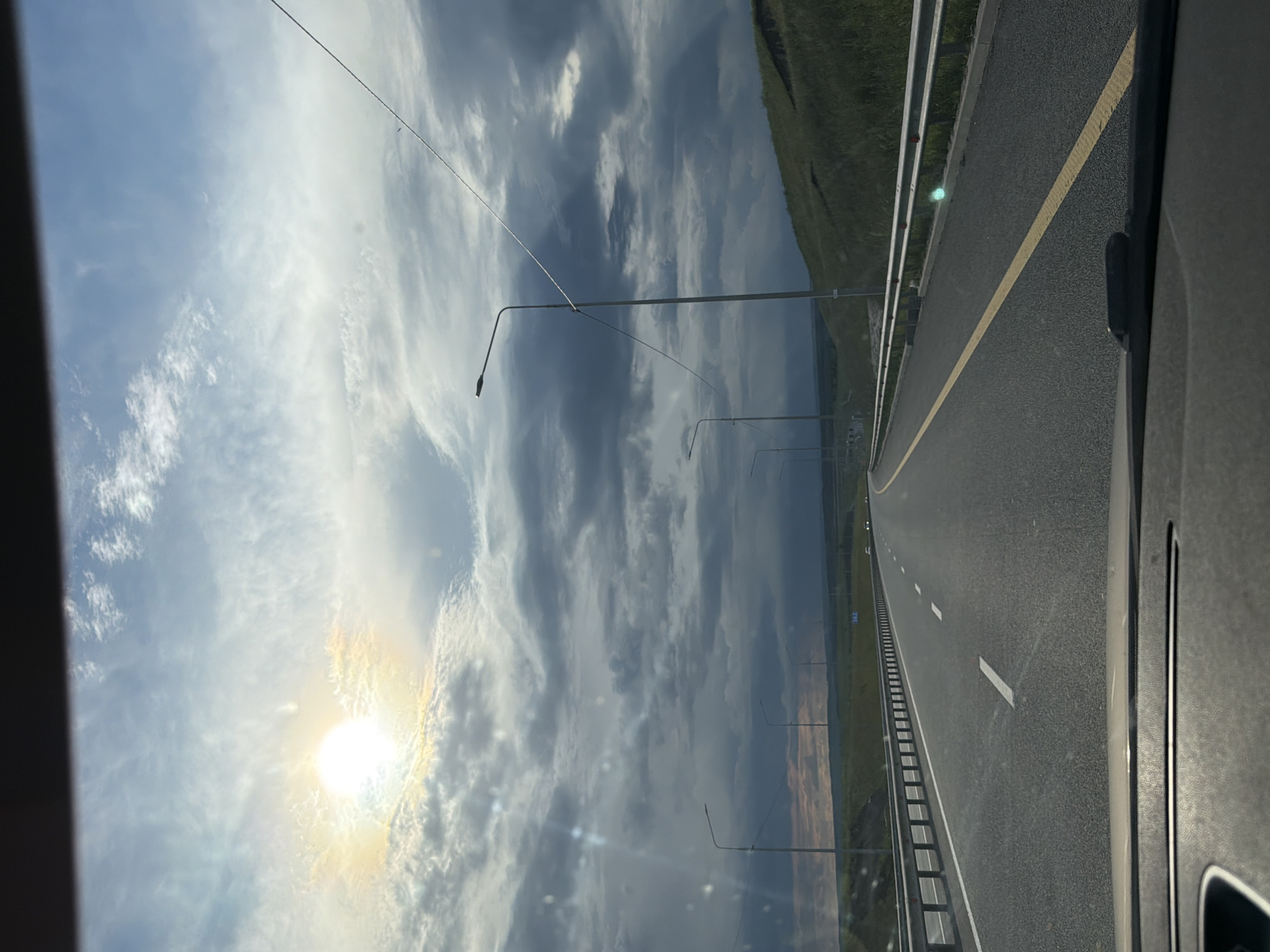 An eye-level, point-of-view shot shows a highway stretching into the distance under a dramatic sky with bright sun and dark clouds. The sun is positioned in the upper left quadrant, casting a bright glow and creating a lens flare effect. Wispy white clouds are visible in the brighter parts of the sky, contrasting with darker, heavier clouds that dominate the upper and middle sections. A yellow line divides the highway, with white dashed lines also present. On the right side of the highway, a grassy embankment rises, dotted with some scattered vegetation. Streetlights line the left side of the road, some with wires extending between them. A metal guardrail runs along the side of the highway. The foreground shows the dashboard of a vehicle. The scene appears to be in a rural or suburban area, with no distinct city features visible.