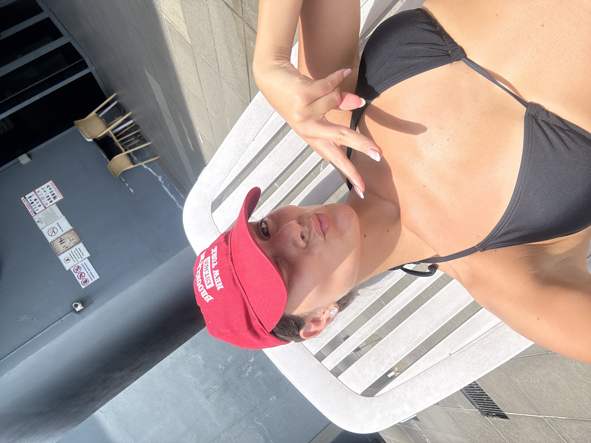 A young woman is depicted relaxing outdoors, likely by a pool, in Kuala Lumpur, Malaysia, under bright daytime sun. She is lying on her back on a white slatted lounge chair, wearing a black bikini top and a red baseball cap. The red cap features white text that reads "BROOKLYN NEW YORK EST. 1830." Her right arm is raised and her fingers are posed in a stylized gesture, highlighting her manicured nails. Her face is turned towards the camera with a slight pout, and she wears stud earrings. The scene is brightly lit, casting strong shadows, indicating clear, sunny weather. In the background, a dark grey wall features a white sign displaying various symbols and text, likely indicating pool rules such as "NO SMOKING" and restrictions like "No Diving" or "No Running." A small security camera or sensor is also visible on this wall. Tiled flooring, often associated with pool areas, is present, along with a dark drain grate. Two light-colored chairs are visible in the distance. The image captures a moment of leisure and sunbathing.