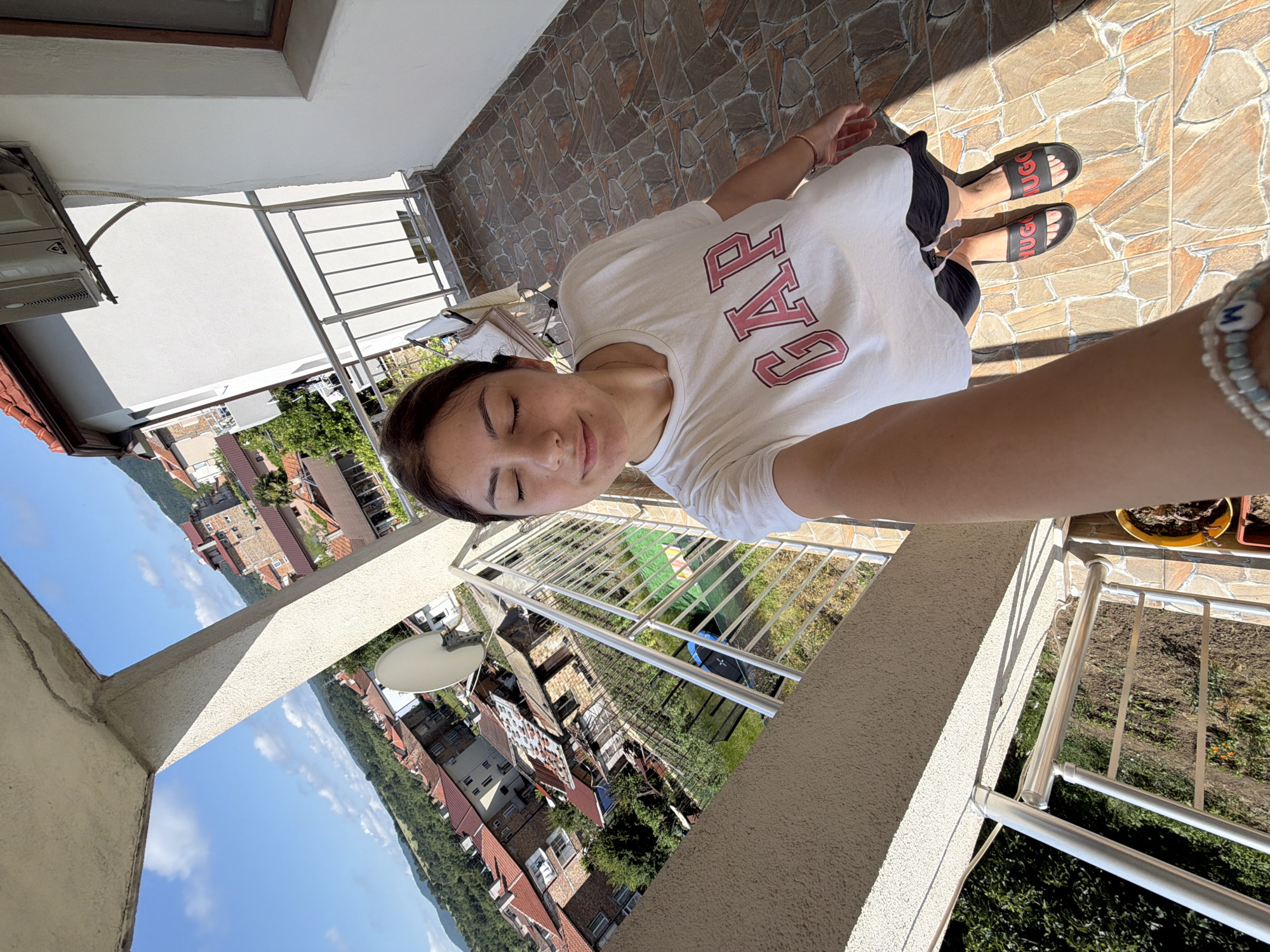 A young woman with dark hair, wearing a white "GAP" t-shirt and black shorts, takes a selfie on a balcony overlooking a town in Tsarevo, Bulgaria. The town features red-tiled roofs and buildings nestled amongst green hills. The balcony has a tiled floor, a metal railing, and a concrete overhang casting a shadow. An air conditioning unit is mounted on the wall of the building. The sky is blue with scattered clouds, and the sun is shining.