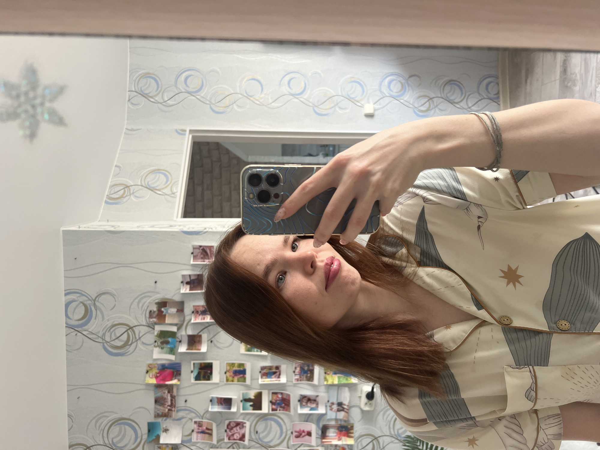 This media file is a mirror selfie taken by a young woman, presented rotated 90 degrees clockwise. The subject is a woman with fair skin, medium-length reddish-brown hair, and light-colored eyes, looking directly into the camera reflected in her phone. Her lips are slightly parted and appear to have a glossy or light pink product. She is wearing what appears to be a light-colored satin or silk pajama top patterned with muted gray, white, and gold abstract shapes, including what looks like stars and foliage. A subtle bracelet is visible on her right wrist. She holds an iPhone with a patterned blue and black case in her right hand, visible in the reflection.
The background, also reflected in the mirror, shows an indoor setting. The walls are covered in light blue wallpaper featuring a repeating pattern of blue, gold, and gray swirls and wave-like lines. To the left, a blurry, iridescent snowflake decal is affixed to a surface. A section of the wall behind the woman is densely decorated with numerous small, square photographs or polaroids arranged in rows, affixed to the wallpaper. In the upper central part of the reflection, a rectangular opening or window reveals a blurry reflection of a brick wall texture. The setting appears to be a personal room, possibly in a residence in Chaikovsky, Russia. There is no visible text in the image, and the time of day or weather conditions cannot be determined.