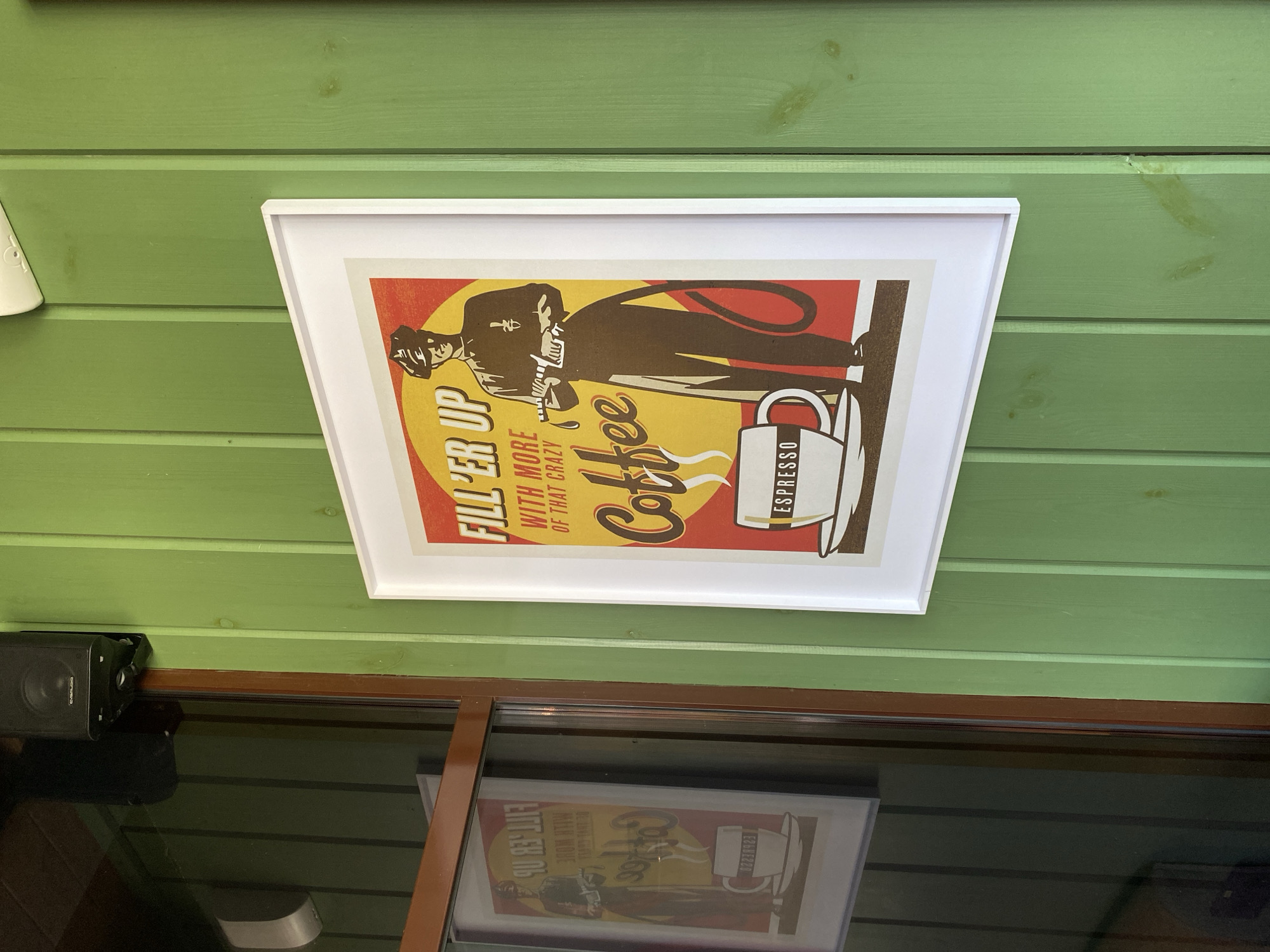 The image shows a framed poster hanging on a green, wood-paneled wall. The poster is a vintage-style advertisement for coffee, featuring a man in a suit holding a fuel hose, presumably to fill up with coffee instead of gasoline. The poster reads "Fill 'er Up with More of That Crazy Coffee ESPRESSO". Below the poster, there's a window reflecting a similar poster, and a black speaker is attached to the wall to the left of the window. The location in Izhevsk, Russia is not evident from the visual content of the image.