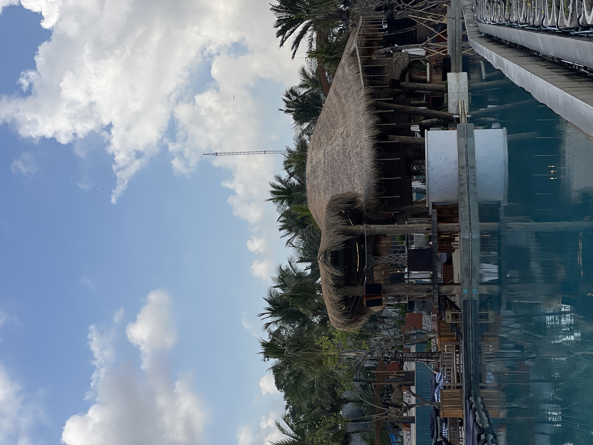 This image captures a resort area in North Kuta, Indonesia, likely during the daytime under a partly cloudy sky. The foreground is dominated by a large, turquoise swimming pool, its surface reflecting the surrounding structures and vegetation. The water is still, indicating a calm environment.
Along the edge of the pool, there are lounge chairs with dark cushions, suggesting a recreational area. Further back, a prominent open-air structure with a thatched roof stands out. This building appears to be a bar or restaurant, featuring wooden pillars, a white circular bar counter, and seating areas. Hanging lights are visible within the structure, and the thatched roof is thick and provides ample shade.
Behind the thatched-roof building, a dense line of palm trees stretches across the mid-ground, characteristic of a tropical location. A tall, slender radio tower is visible in the distance, piercing the sky above the trees. The sky itself is a pale blue, dotted with fluffy white and gray clouds.
The overall impression is of a serene and upscale tropical resort, designed for relaxation and leisure, with a distinct Indonesian architectural influence. There are no people visible in the image.