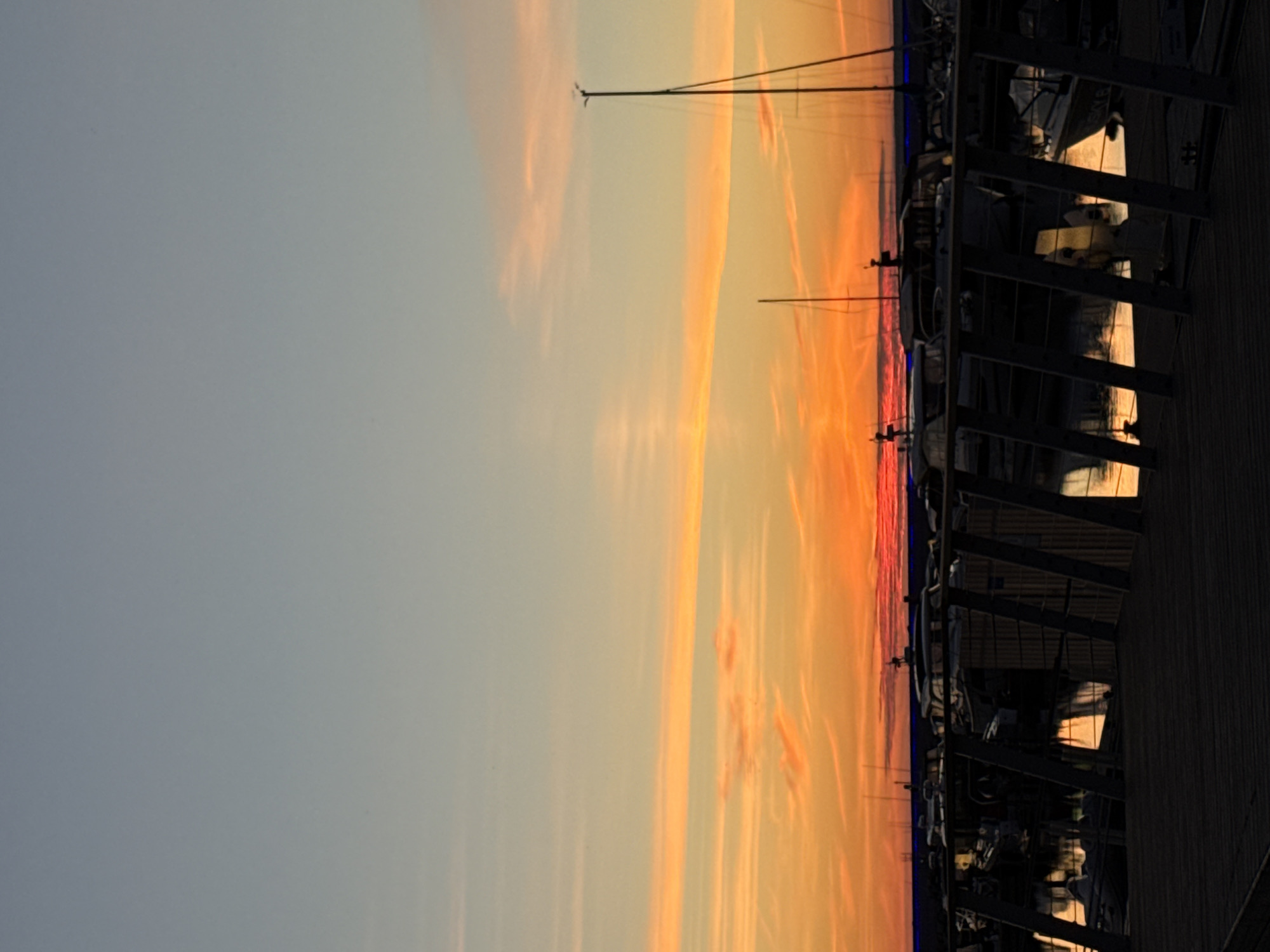 The image depicts a marina at sunset, likely located in the city of Tallinn, Estonia. In the foreground, a dark wooden pier or boardwalk dominates the lower portion of the frame, with railings extending across it. Beyond the pier, a body of water reflects the vibrant colors of the sunset. Several sailboats and motorboats are moored in the marina, their masts and silhouettes silhouetted against the colorful sky. The sky itself transitions from a pale blue at the top to a fiery orange and yellow near the horizon, with wispy pink clouds streaking across. The overall atmosphere is one of calm and serenity as the day draws to a close. There are no people visible in the image.
