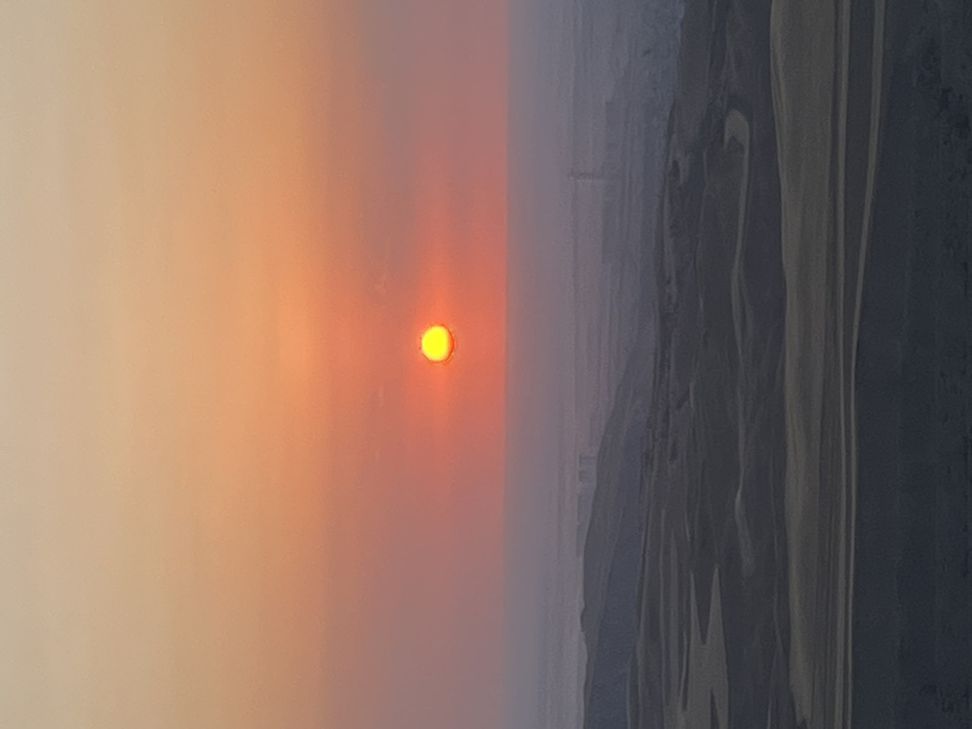 The image depicts a vast, hazy desert landscape at sunset. The sun, a bright orange orb, is centered in the upper half of the frame, casting a warm glow across the sky which transitions from a pale yellow at the top to orange and then pink towards the horizon. In the middle ground, a faint layer of haze obscures distant elements, but hints of structures or a city are visible. The foreground is dominated by undulating sand dunes with dark, shadowed curves, creating a textured and layered appearance. The overall impression is one of a serene and expansive desert environment during twilight. There are no people or animals visible in the image. The location is identified as Besh Kungei, Kyrgyzstan.