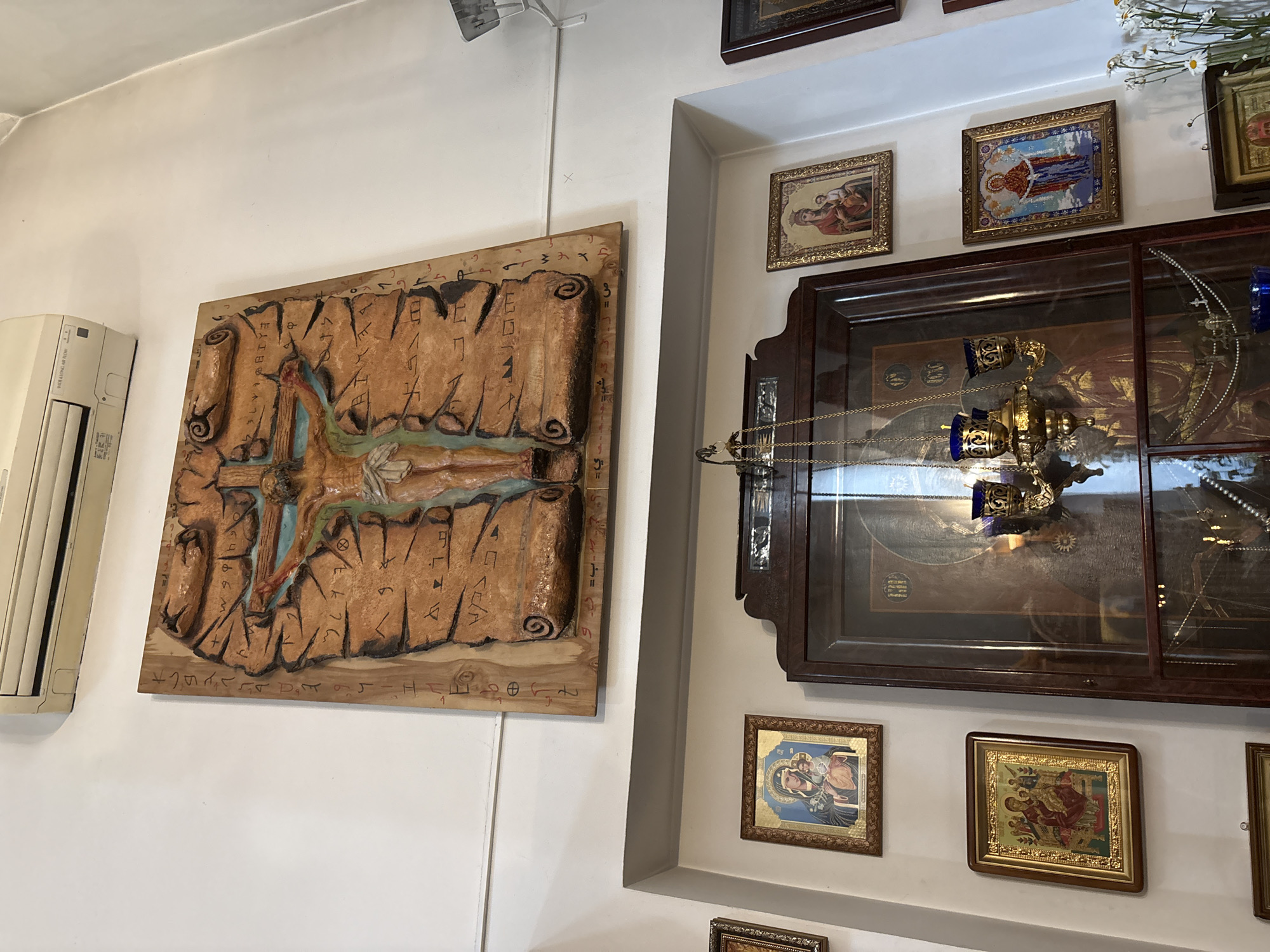 This image shows a wall in what appears to be a religious building, possibly a church or chapel, located in Sestroretsk, Russia.
Dominating the upper portion of the image is a large, relief carving on wood depicting the crucifixion of Jesus Christ. The carving features Jesus on the cross, with scrolled parchment-like textures surrounding him, inscribed with what appear to be religious symbols or script. A portion of an air conditioning unit is visible on the white wall above the carving.
Below this central piece, several framed religious icons are displayed on the wall. To the left, there are two icons, the upper one showing the Virgin Mary holding the infant Jesus, and the lower one depicting a similar scene with multiple figures. To the right, two more icons are visible. The upper right icon also features the Virgin Mary and child, while the lower right icon shows a woman in prayer or adoration.
In the center of the lower half of the image, a large, ornate wooden cabinet with glass doors is present. Inside, a hanging religious lamp (likely an icon lamp or censer) with blue glass elements is visible. The cabinet contains other religious artifacts, including what appears to be a large icon or painting behind the lamp. A string of pearls or rosary beads is also visible within the cabinet.
To the right of the cabinet, a vase with white flowers, possibly daisies, is placed on a ledge. Another framed icon is partially visible below the flowers.
The environment is dimly lit, with the light source appearing to come from above and from within the cabinet. The walls are white, and the overall impression is one of a sacred space adorned with religious art.
