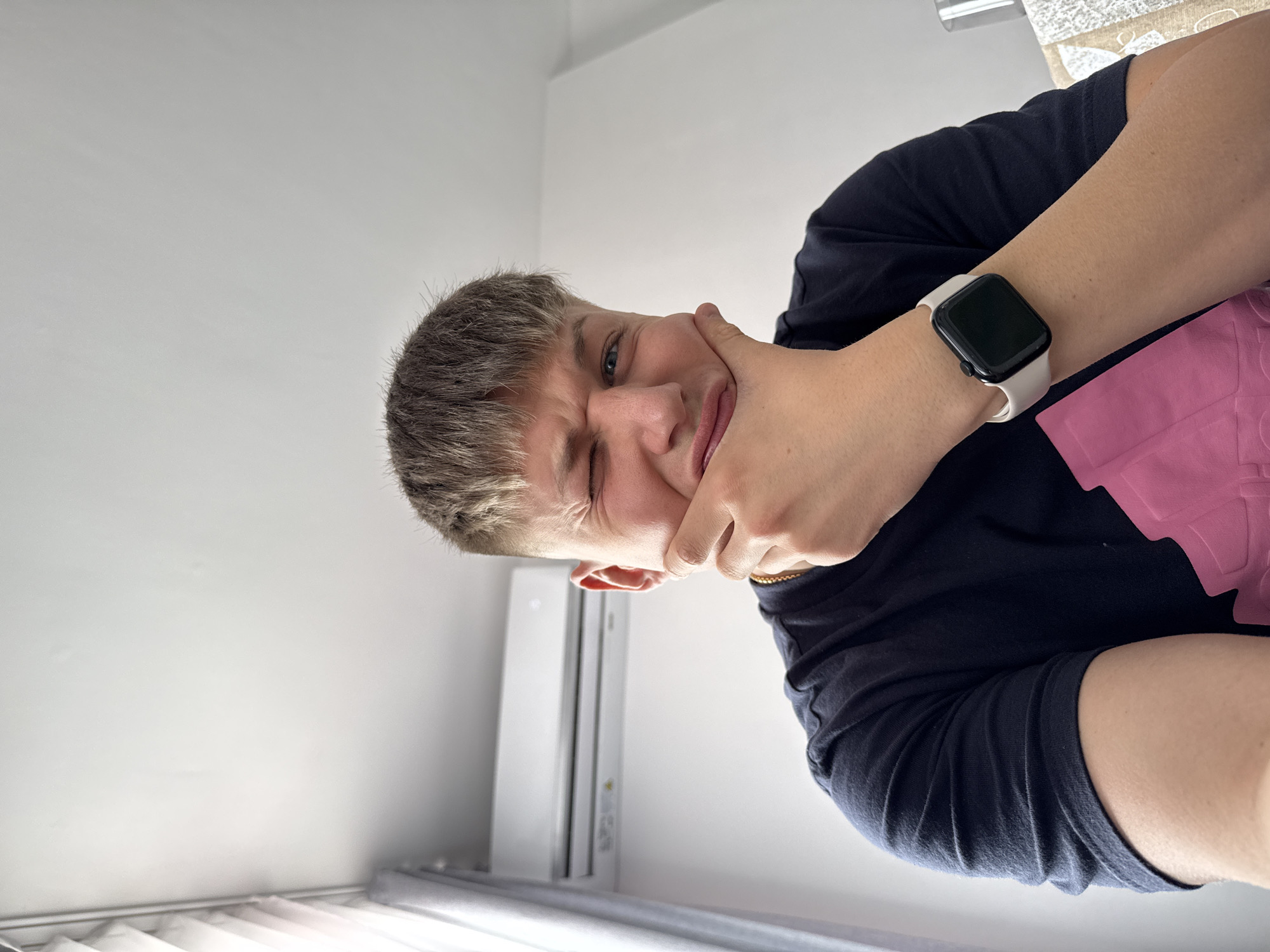 A young person, cropped closely, is holding their cheek with their hand, winking at the camera. The person has short, light brown hair. They are wearing a dark blue t-shirt and a white smartwatch. In the background, a white air conditioning unit is visible, and partially seen window blinds at the bottom of the frame. The scene appears to be indoors. The exact location within Plovdiv, Bulgaria is not discernible.