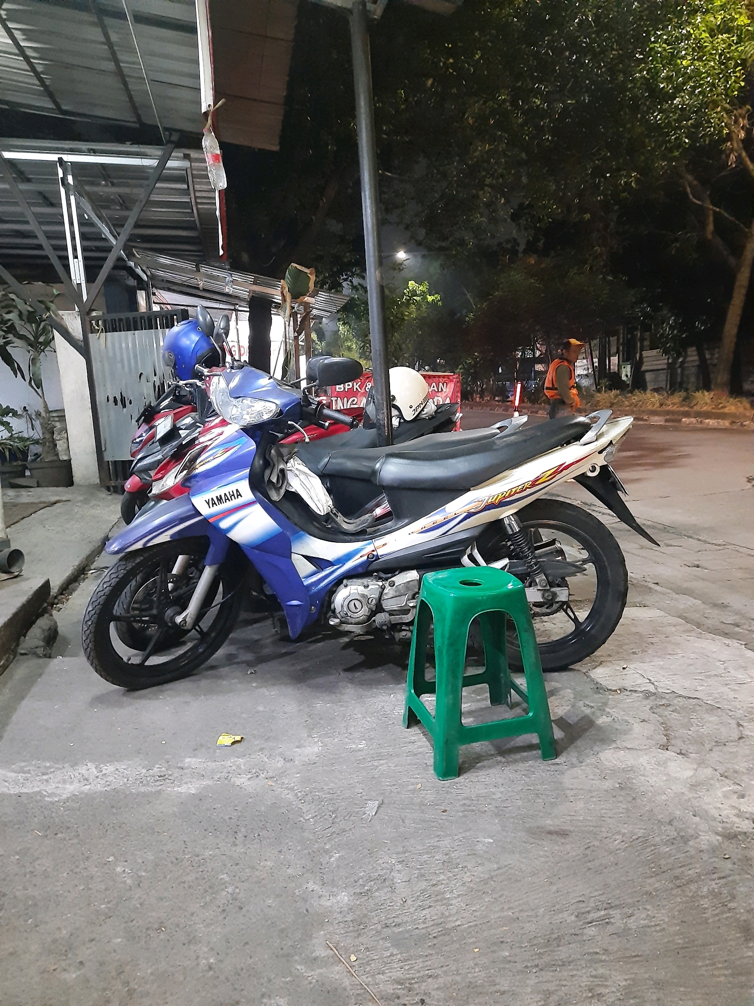 A night-time street scene in Bandung City, Indonesia, features two parked motorcycles, one blue and white, the other red. A green plastic stool sits in front of the blue and white motorcycle. In the background, a person wearing an orange vest is visible on the sidewalk. Trees and streetlights are present. A plastic bottle hangs from a string above the motorcycles.