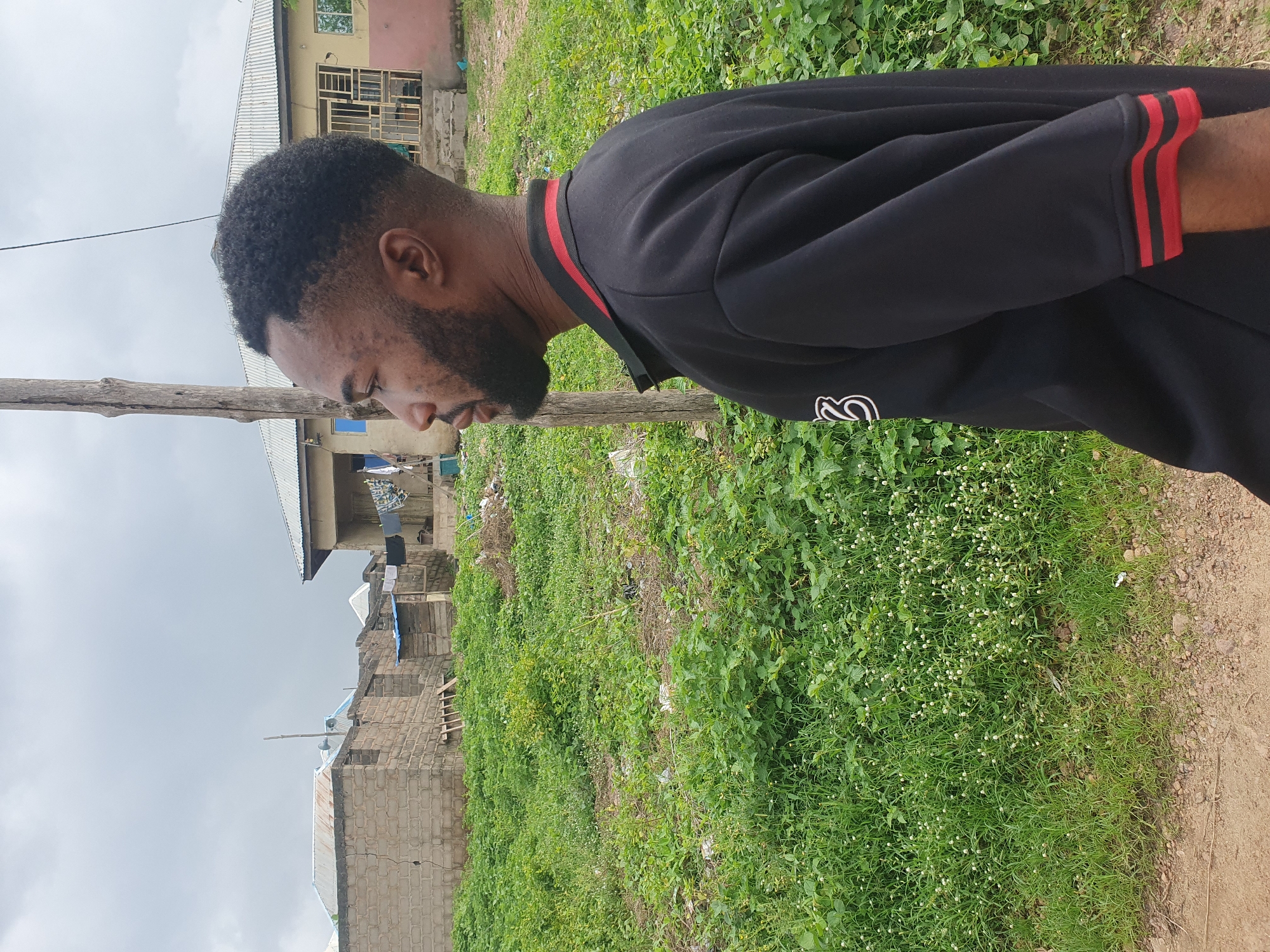 An outdoor, medium shot shows a man from the chest up, in profile facing left, standing in a grassy, undeveloped area in Lokoja, Nigeria. The man is wearing a black polo shirt with red trim on the collar and sleeves, and has a neatly trimmed beard and short, curly hair. Behind him, to the left, is a partially constructed brick building. To the right and center, there are a few houses with corrugated metal roofs, and laundry is hanging on a clothesline. The ground is covered in green grass and some small white flowers, with a dirt path visible in the foreground. The sky is overcast.