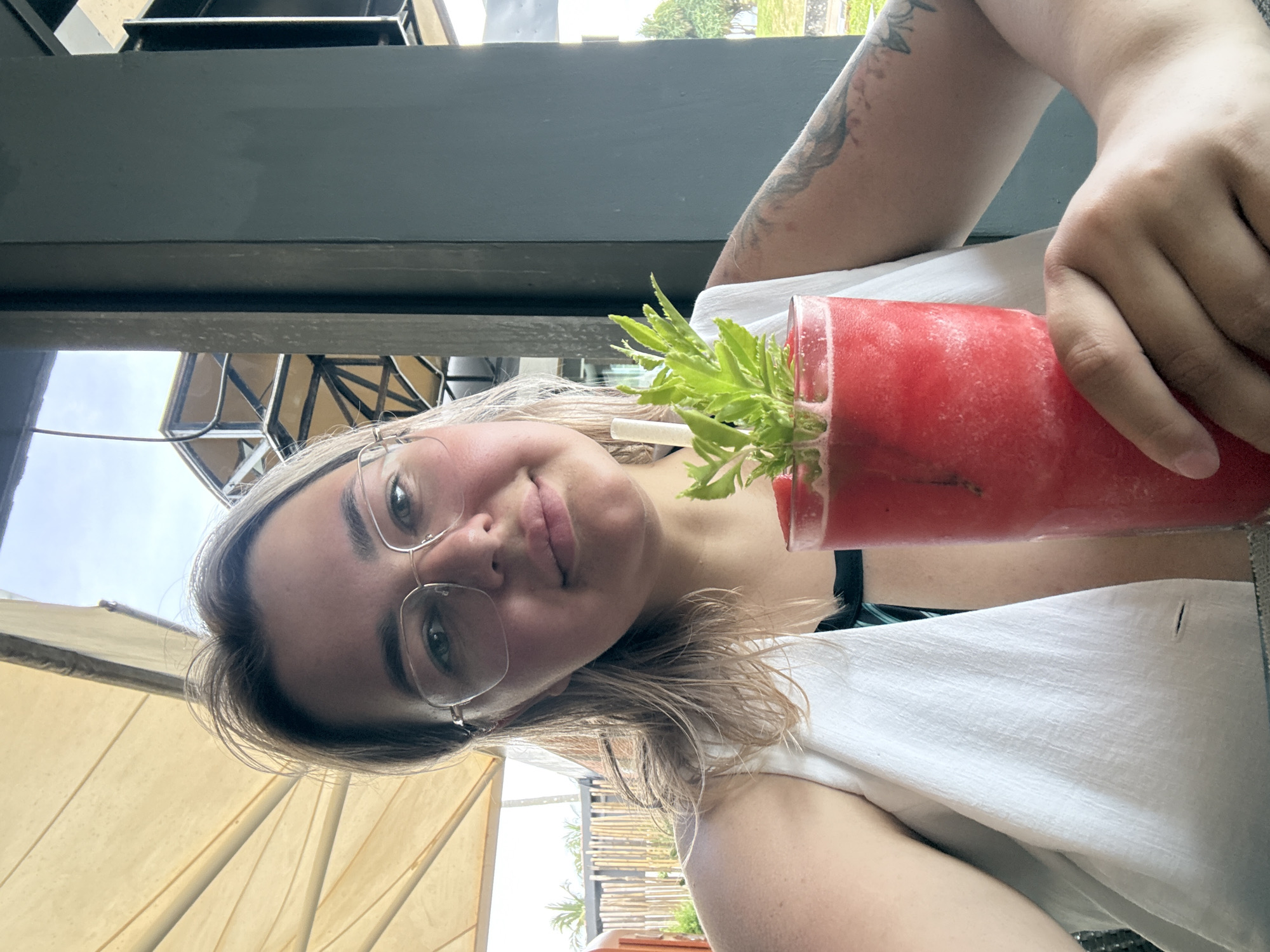 A young Caucasian woman with blonde hair, wearing glasses and a white vest over a dark swimsuit, is taking a selfie. She is holding a tall glass of red, slushy beverage with a green sprig and a straw. She is seated outdoors at a table, possibly at a restaurant or cafe, with a thatched umbrella structure overhead and a dark pillar to her right. In the background, there are bamboo fences, foliage, and a glimpse of the sky. The overall impression is of a relaxed, sunny day.