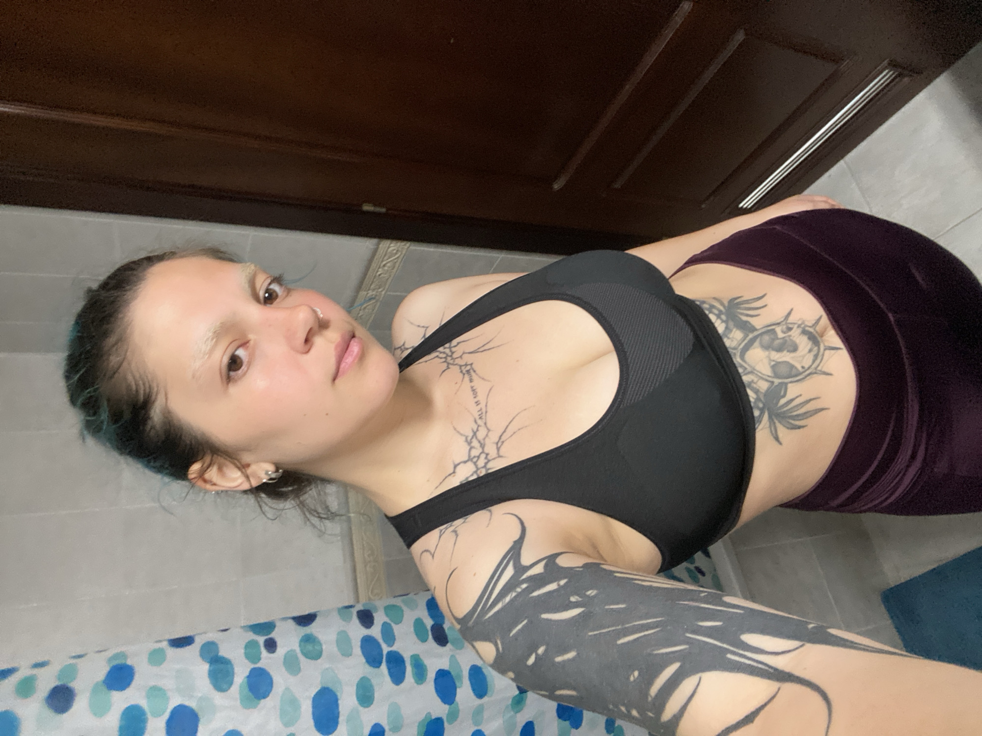 A young woman with dark hair, light eyebrows, and visible tattoos on her left arm, chest, and abdomen is standing in a bathroom. She is wearing a black sports bra and dark purple leggings. A blue and white polka dot shower curtain is visible to her left, and a dark brown door is on her right. The background appears to be a tiled wall. The image seems to be a selfie taken by the woman.