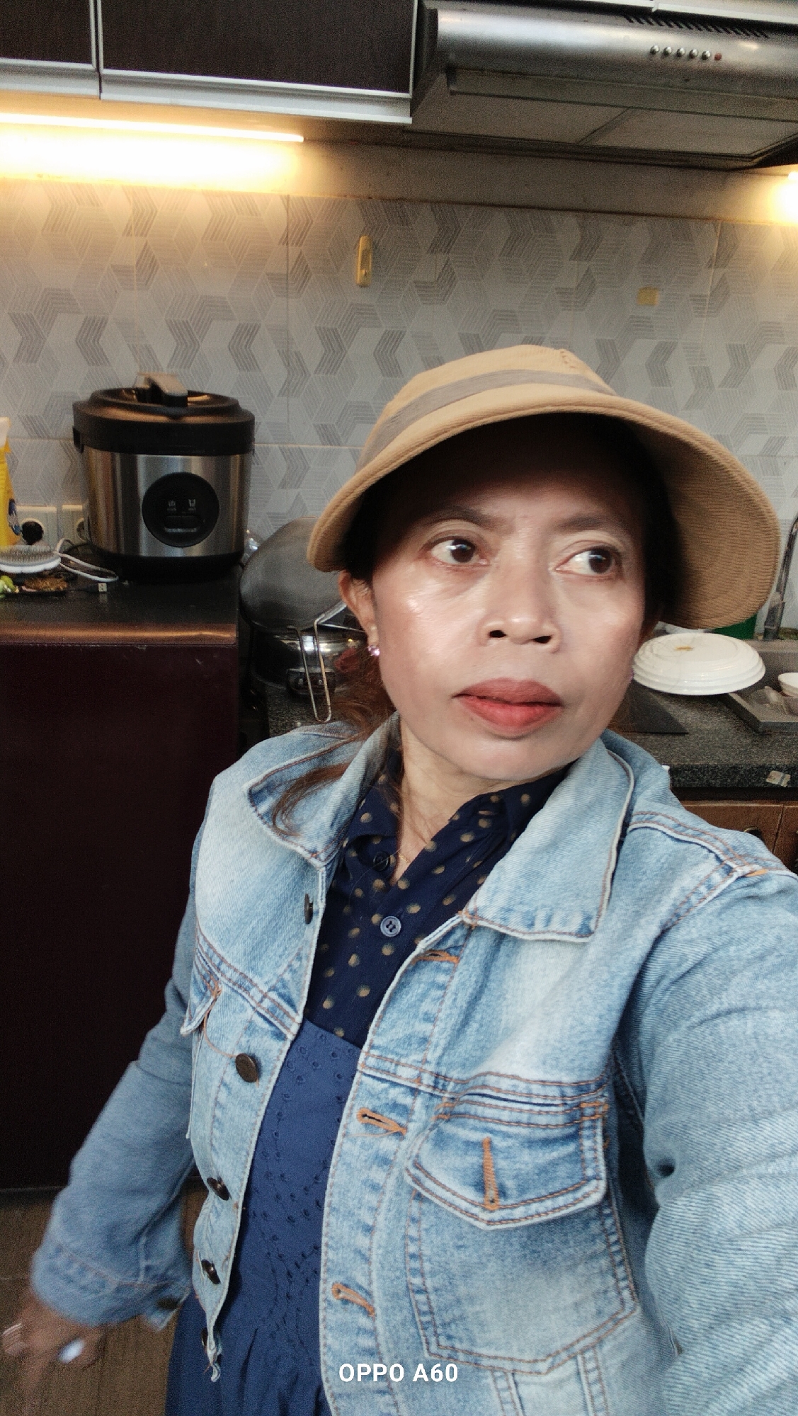 A medium shot captures an Indonesian woman in a dimly lit kitchen, wearing a light brown sun hat, a blue polka dot shirt, and a denim jacket. She is looking away from the camera to her right. The background shows kitchen cabinets with strip lighting, a patterned backsplash, and various kitchen appliances including a rice cooker. The overall scene appears to be in North Kuta, Indonesia.