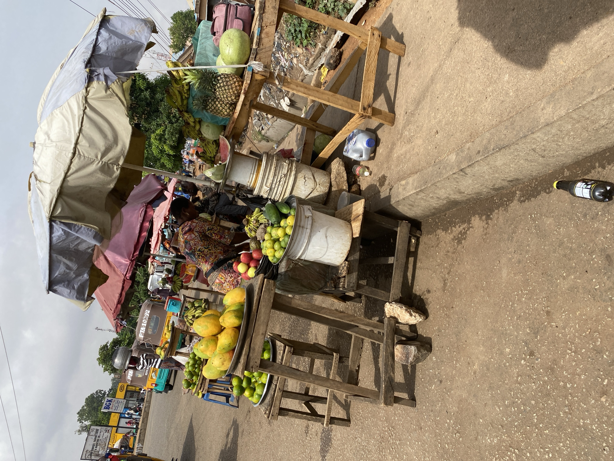 The image depicts a roadside fruit stand in Bukuru, Nigeria. A vendor sits at the stand under an umbrella. The stand displays mangoes, limes, bananas, apples, pineapples, and watermelons. The stand is constructed of wooden planks and is set up on a concrete walkway alongside a busy road with auto rickshaws and other vehicles. A discarded plastic bottle sits near the walkway’s edge. In the background, there is signage indicating businesses or advertisements and other vendors.