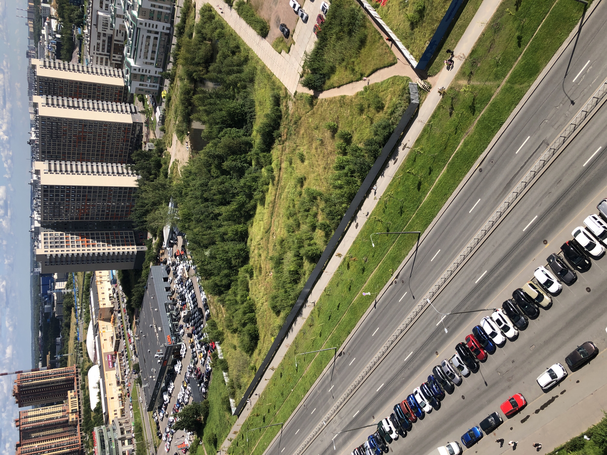 This is a high-angle aerial shot of a city street and surrounding buildings. In the foreground, a multi-lane road runs diagonally from the bottom left to the top right. Several cars are parked along the right side of the road. A grassy median with a sidewalk and streetlights separates the road from a fenced-off green space. Beyond the green space, a large building complex with a dark roof and many parked cars is visible, with a small body of water and trees to the right of it. In the background, numerous tall residential buildings dominate the skyline, interspersed with some lower-rise structures and distant cityscapes under a partly cloudy sky.