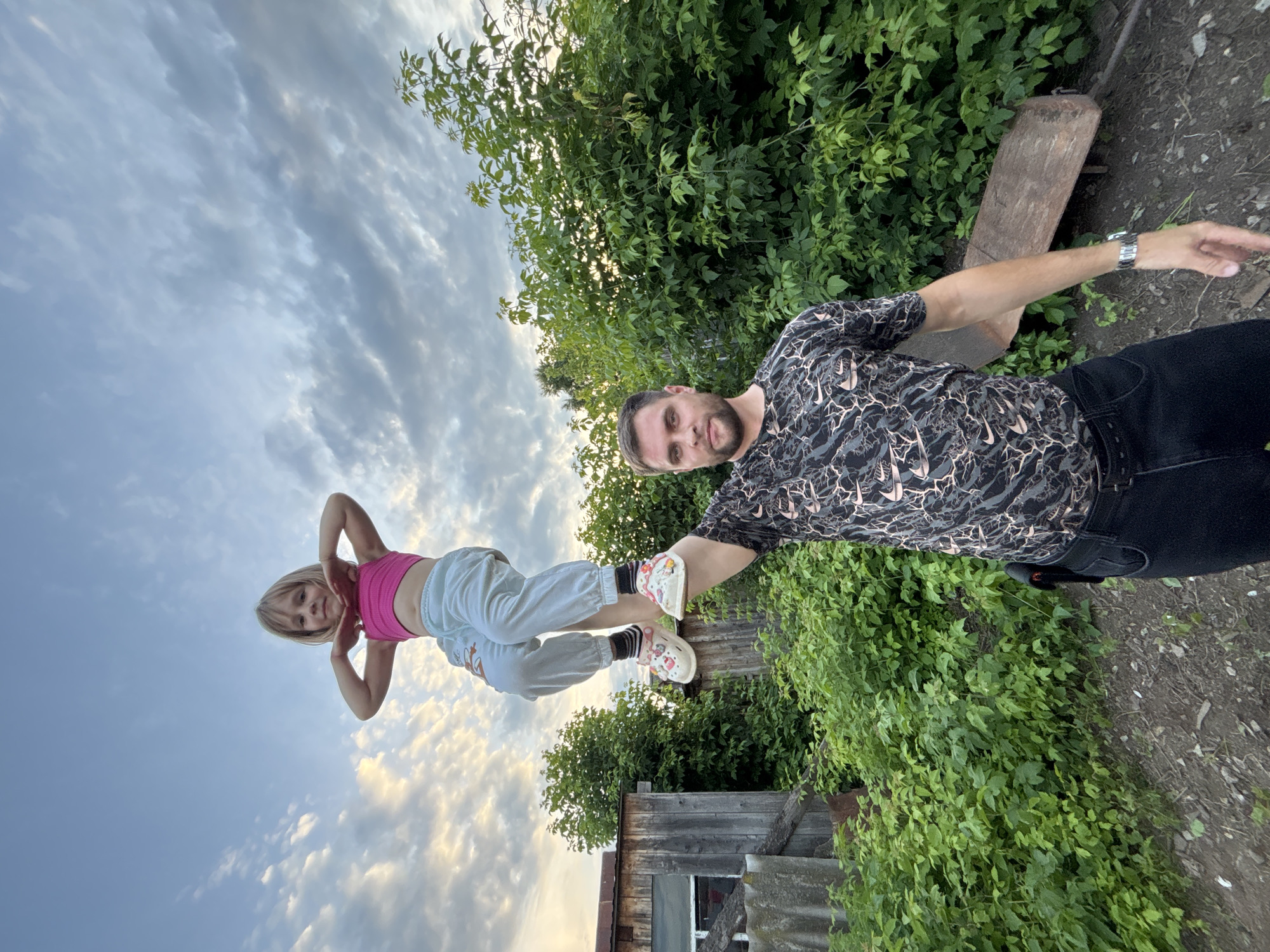 In an outdoor scene set in Маркидоновка, Russia, a man and a young girl are captured in a dynamic pose against a backdrop of lush greenery and a cloudy sky. The man, wearing a black patterned t-shirt and dark pants, extends his right arm, his hand pointing forward. He wears a watch on his left wrist. The girl, positioned in front of him, is balancing on what appears to be a wooden fence or railing. She is wearing a pink sports bra, light-colored pants, and distinctive white clogs with colorful designs. Her arms are raised above her head, hands near her face.
The low angle of the shot emphasizes the sky, which is filled with scattered clouds, suggesting it is either early morning or late afternoon. The surrounding environment is characterized by dense green foliage and hints of wooden structures, indicative of a rural or suburban setting. The overall mood appears playful and energetic, with the two subjects engaged in an activity that showcases balance and perhaps trust. There is no visible text in the image.