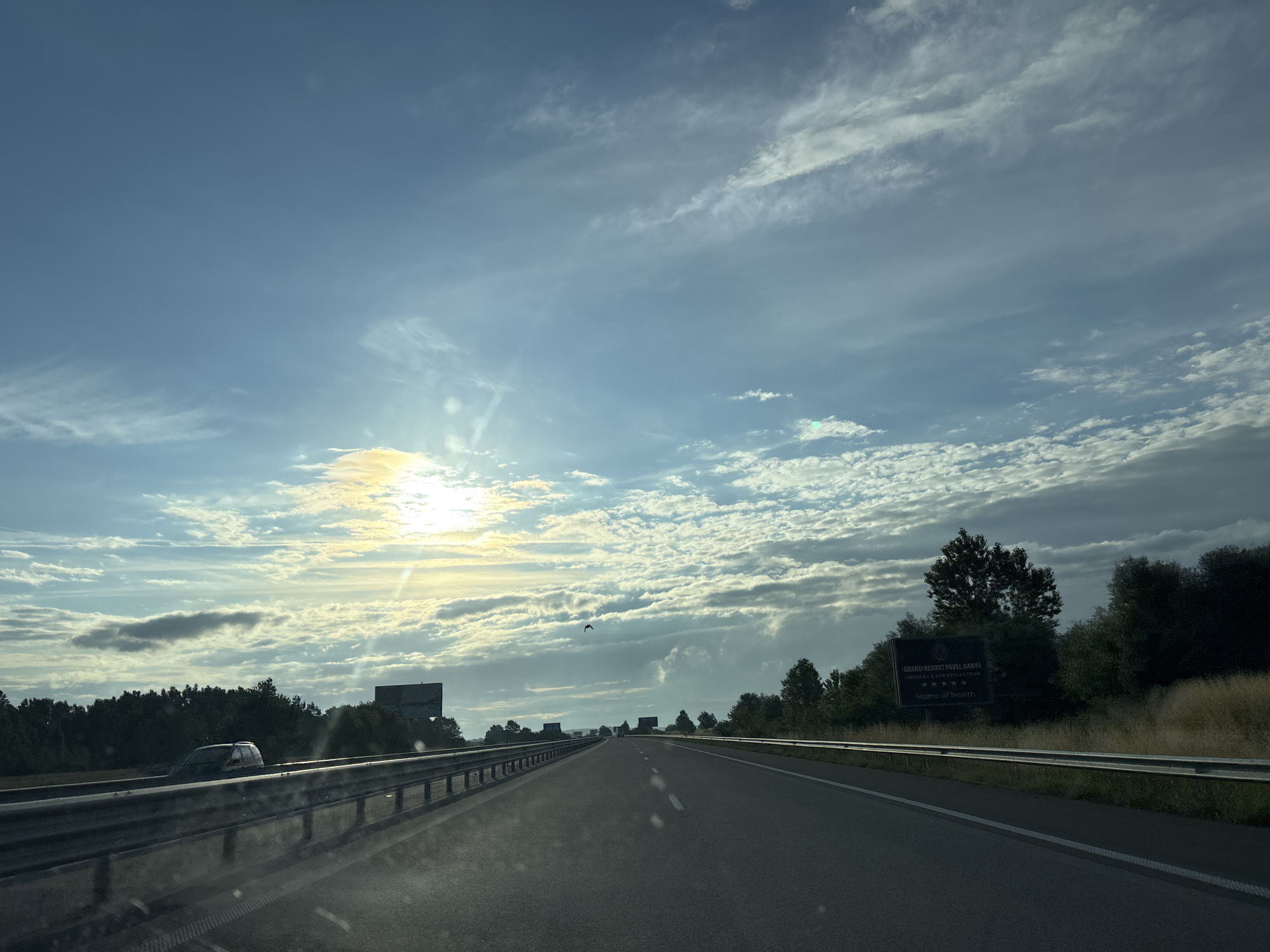An eye-level, wide-angle shot captures a view from a vehicle traveling on a highway. The sun is visible in the upper left quadrant, casting a bright glare. The sky is blue with scattered white clouds and streaks of cirrus clouds. The road ahead stretches into the distance, with guardrails on both sides. Trees line the sides of the highway, and a sign that reads "GRAND RESORT PAVEL BANYA" with five stars and "HOME OF HEALTH" is visible on the right. The location is the highway approaching Kaloyanovets, Bulgaria. No people are visible in the shot.